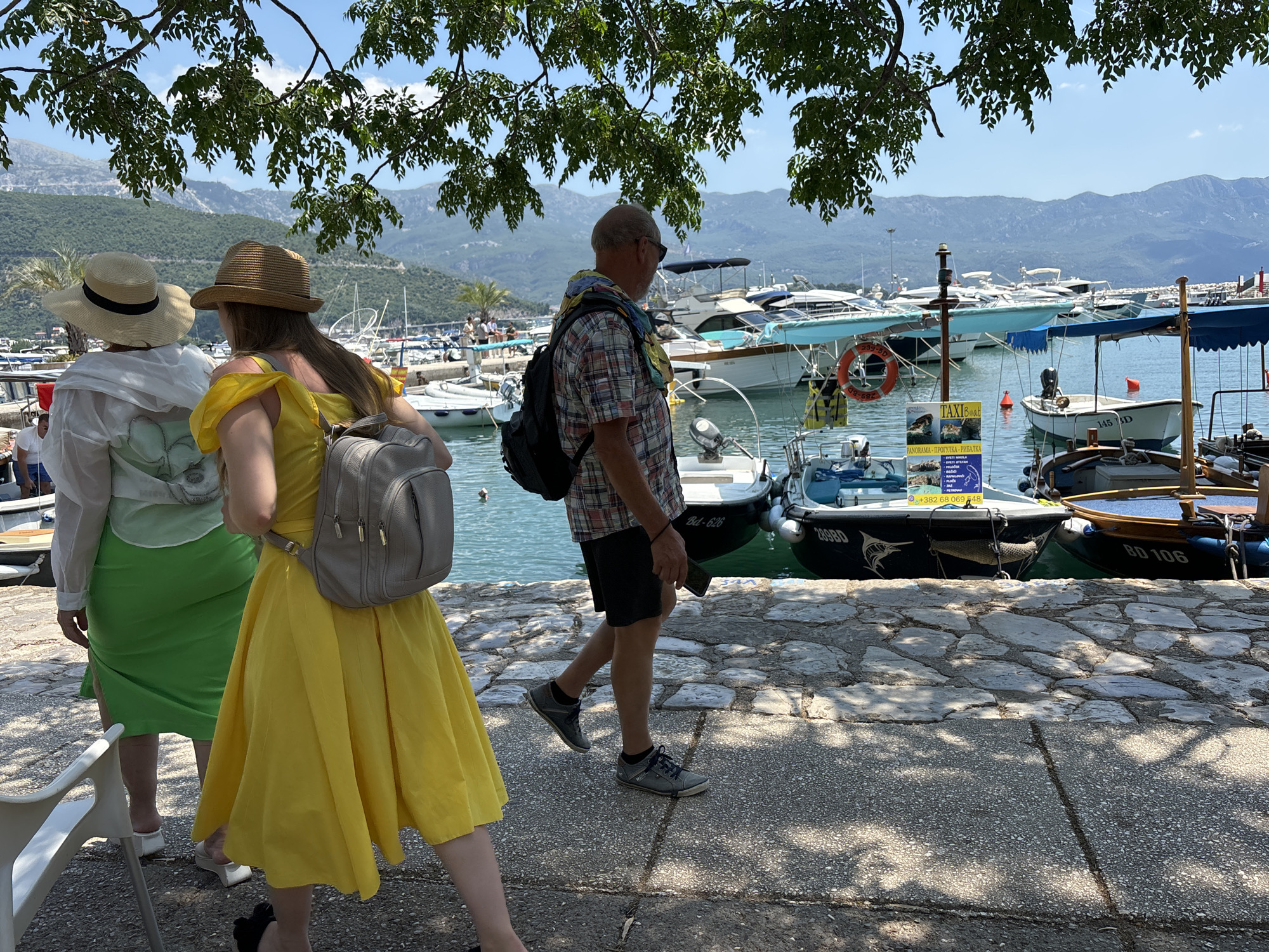 This image depicts a view of the waterfront promenade in Budva, Montenegro, on a sunny day. The foreground shows a cobblestone pathway with shadows from overhead trees. To the left, two women are walking away from the camera. The woman on the left wears a wide-brimmed straw hat, a white shirt, and a green skirt. The woman on the right is wearing a straw hat, a yellow dress, and a grey backpack. A man in a plaid shirt and dark shorts is walking away from the camera in the middle ground, also with a backpack. In the background, the clear blue water of the Adriatic Sea is visible, filled with numerous docked boats and yachts of various sizes. Beyond the harbor, a lush, green mountainous landscape stretches across the horizon under a bright blue sky. The overall scene conveys a relaxed, summer atmosphere at a popular coastal tourist destination.