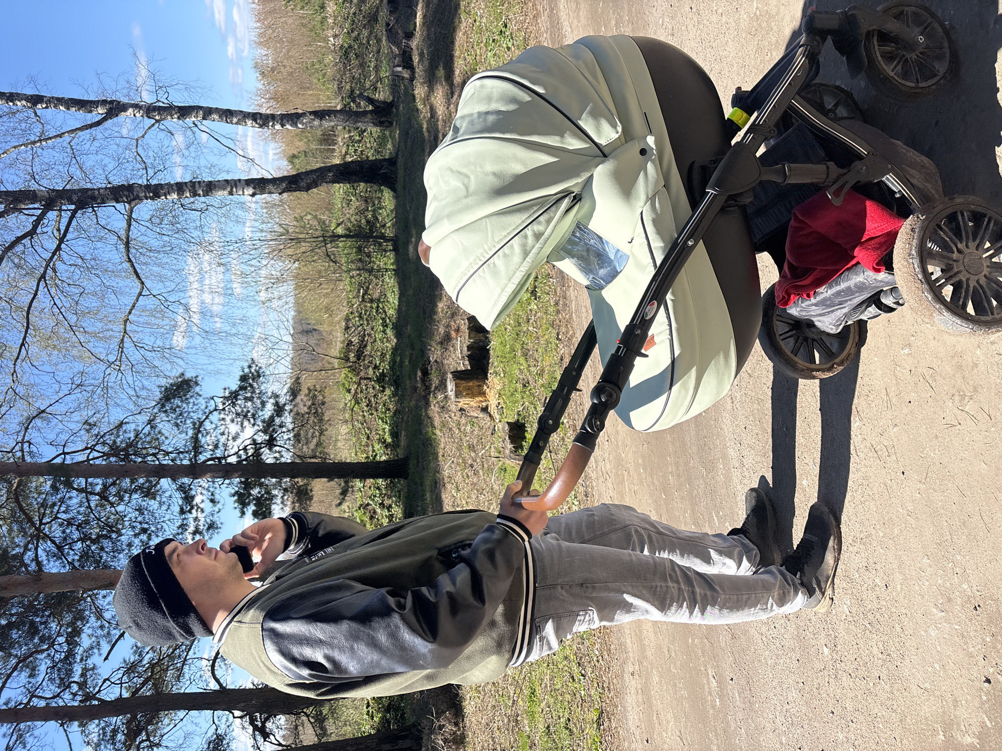A man in a black beanie, green and black jacket, and grey jeans stands on a dirt path in Раздолье, Russia. He holds the handle of a stroller with a light green bassinet while talking on his mobile phone. The stroller has mud-covered wheels and a red cloth stowed underneath. Trees are visible in the background under a blue sky.