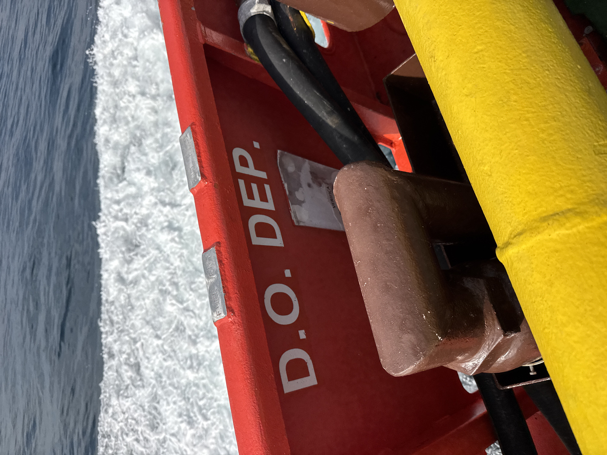 This is a close-up, eye-level shot of the stern of a boat, likely a working vessel, in the waters off Marina di Ravenna, Italy. The focus is on the red-painted railing and a portion of the hull. White foam from the boat's wake is visible in the background, with the blue water stretching to the horizon. On the red railing, the white letters "D.O. DEP." are prominently displayed. To the left, a silver metal plate with faint lettering is affixed to the railing. To the right, a thick black hose is secured to the railing. A brown, pipe-like fixture protrudes from the deck. A bright yellow pipe runs diagonally across the lower portion of the frame. The scene appears to be part of the operational equipment of a vessel, suggesting industrial or commercial maritime activity in the area.