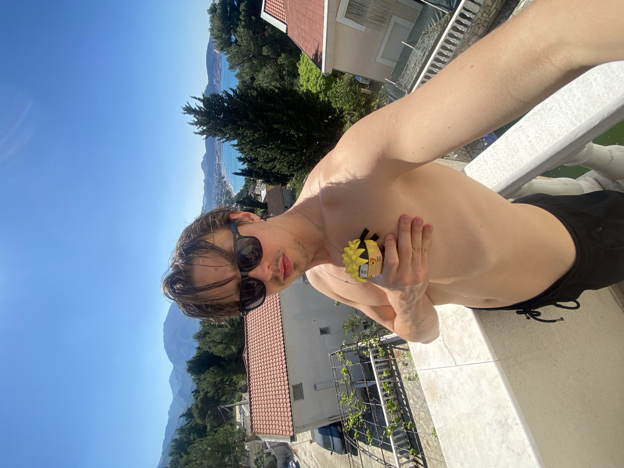 A shirtless man in sunglasses and black shorts stands on a white balcony in Šušanj, Montenegro, holding a Naruto figurine. Behind him, a residential area with red-tiled roofs and lush greenery slopes toward the Adriatic Sea, under a clear blue sky. Mountains are visible in the distance.