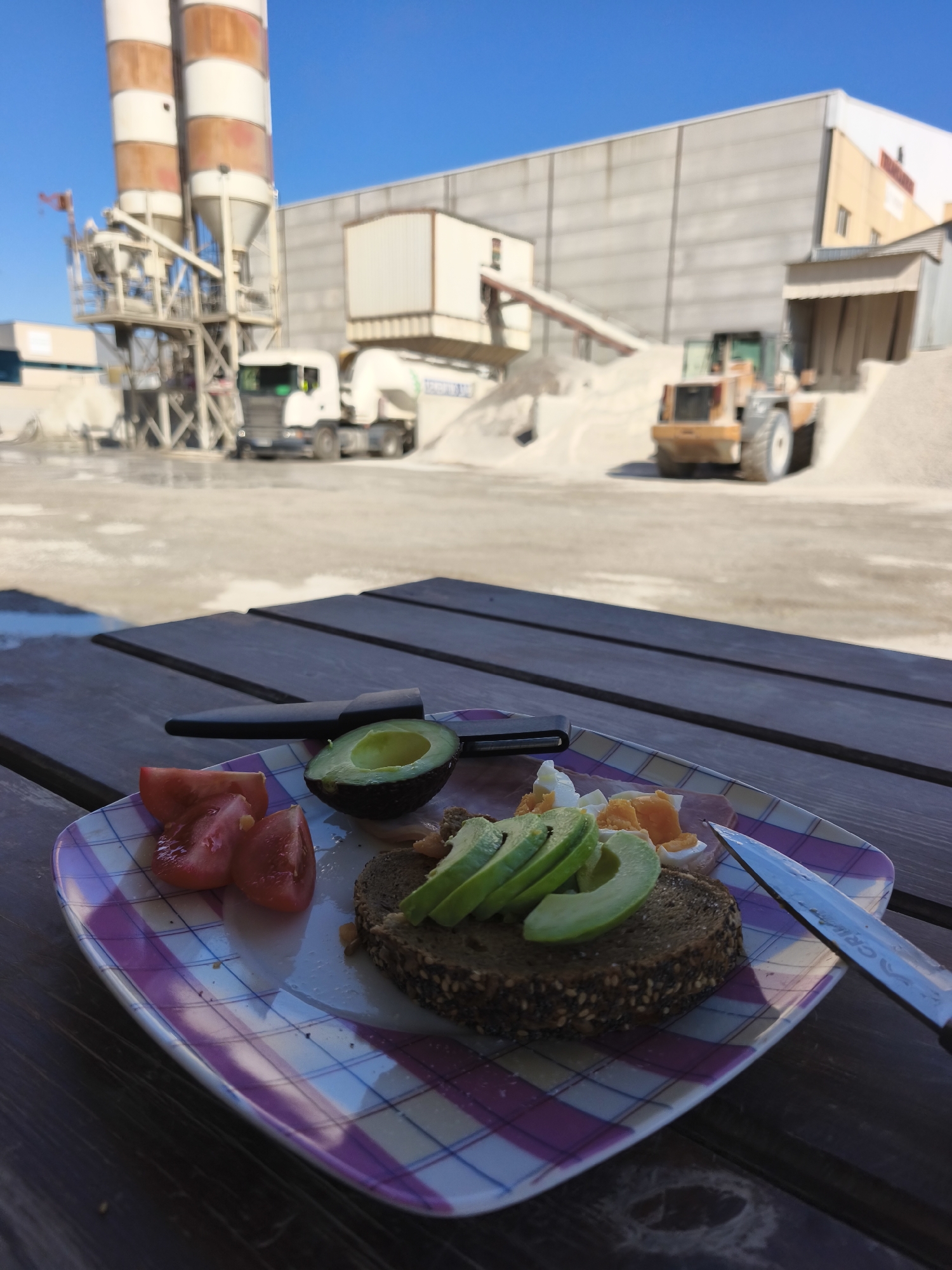 The image presents a plate of food on a wooden picnic table, with an industrial backdrop. The plate contains sliced tomatoes, half an avocado, sliced avocado on a piece of dark bread, and chopped eggs and ham. A knife and a peeler are also on the plate. The background shows a concrete plant or quarry, featuring silos, trucks, and heavy machinery. The scene is set in the city of Polígon Industrial Dels Vents, Spain, indicated by the type of industrial structures present. The sky is clear and blue, suggesting a sunny day. No people are visible.