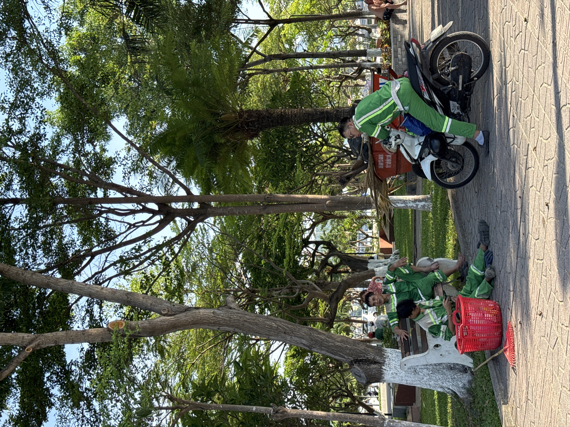 In a park setting in Nha Trang Ward, Vietnam, three individuals in matching green and white work uniforms are taking a break. One person is seated on a bench, while another is sitting on the ground beside a red plastic basket and a red rake. The third person is seated on a white motorcycle. The ground is paved with textured tiles, and a sidewalk is visible. Lush green trees and foliage surround the scene, with a blue sky overhead. In the background, a vehicle and other people can be seen, suggesting a public space. The overall impression is one of a warm, sunny day, with workers pausing their tasks.