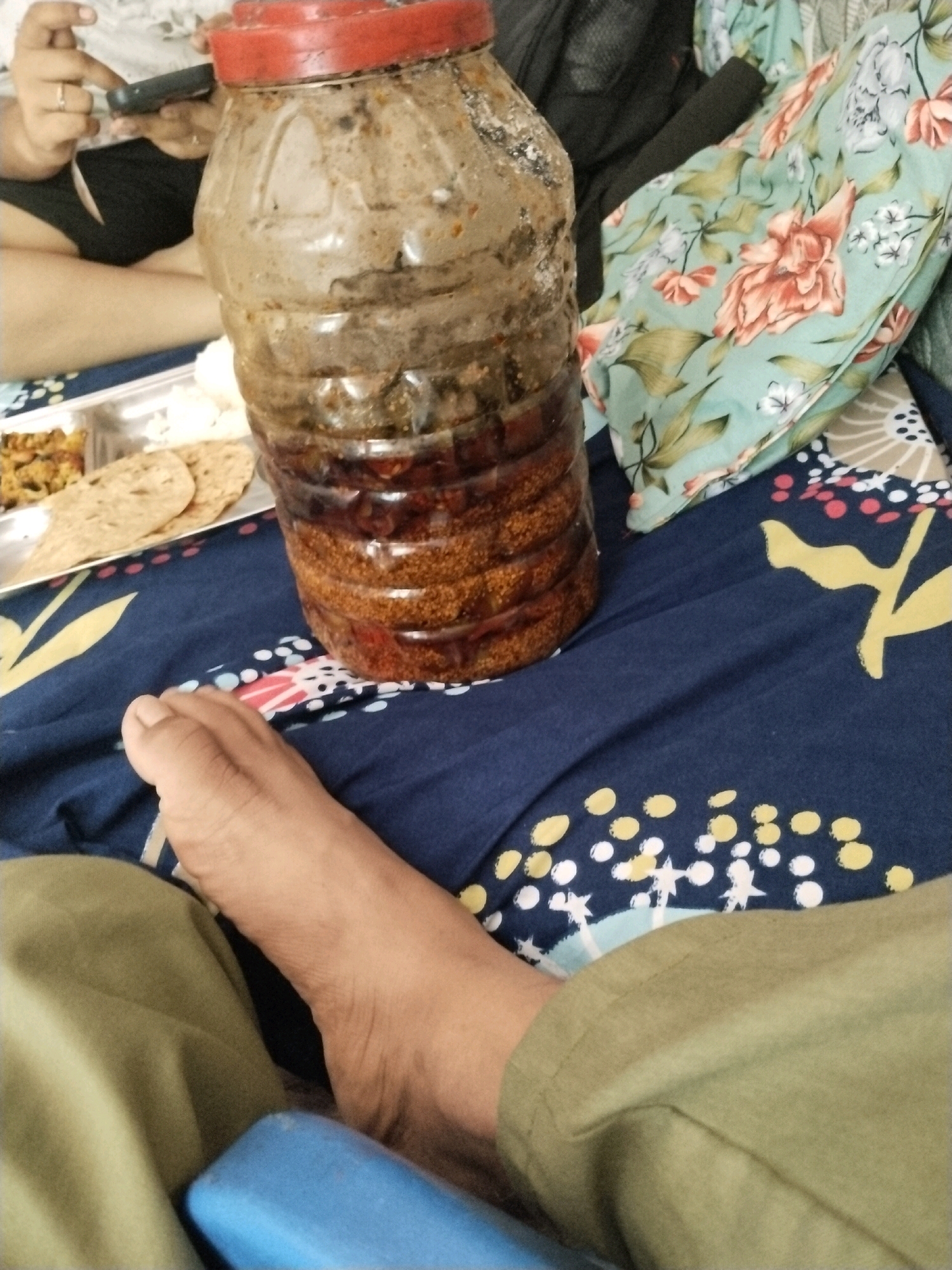 The image displays a close-up, overhead view of a person's foot and legs resting on a dark blue fabric with a floral pattern. To the right of the foot is a large, clear plastic bottle filled with a dark, layered food item, possibly pickles, with a red lid. The bottle is positioned on the floral fabric. In the background, on a silver plate, there appears to be food, including flatbreads and a dish with diced vegetables, partially obscured by the bottle. To the left, a person's hands are visible, holding a mobile phone and wearing rings on two fingers. The overall setting appears to be a domestic environment, possibly a bed or sofa, in Bilaspur, India.