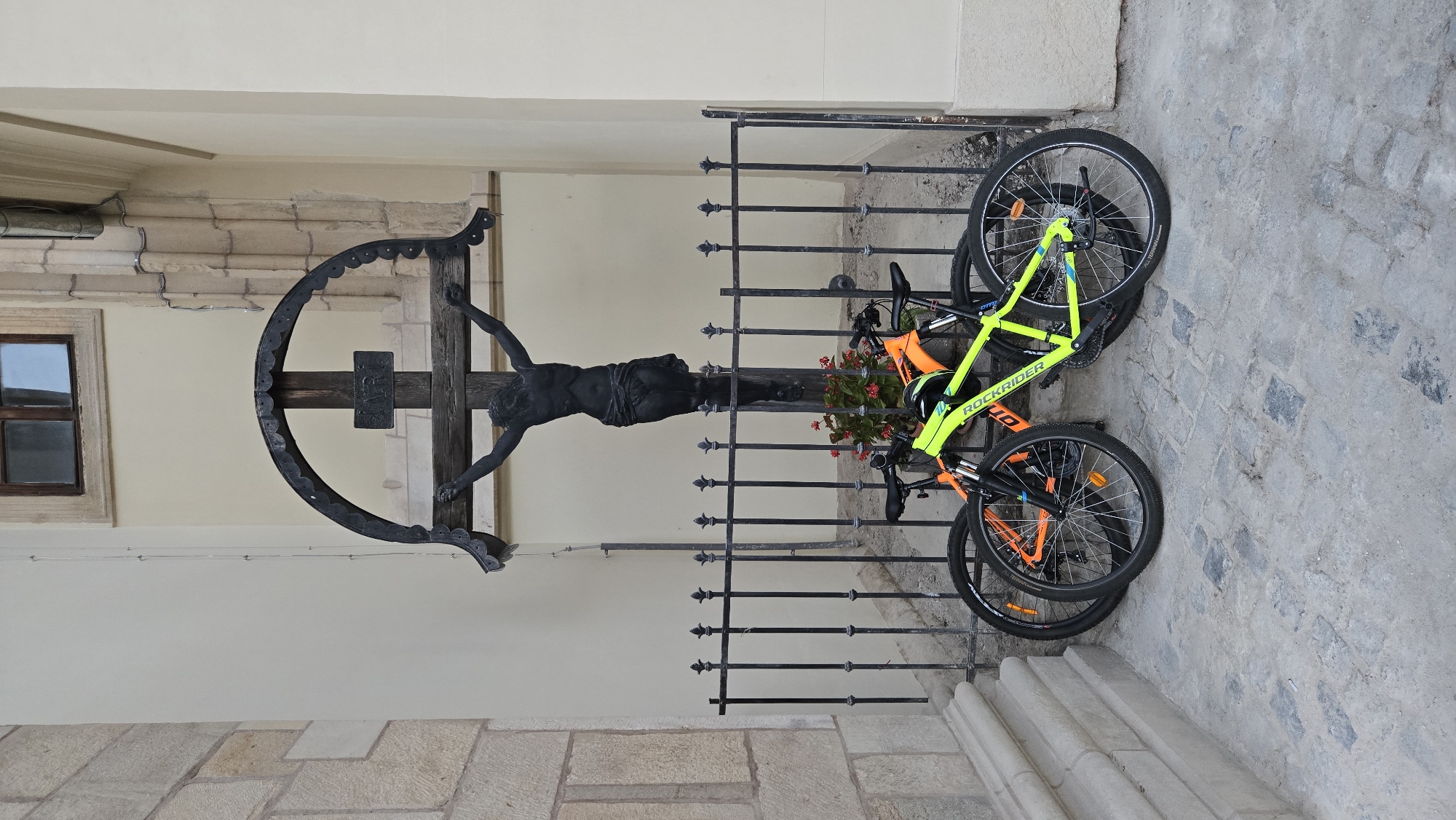 In Cluj-Napoca, Romania, at the Saint Michael's Church, a black statue of Jesus on the cross stands behind a metal fence. Two bicycles, one bright yellow and the other orange, are parked in front of the fence. The statue is positioned centrally in the frame, with the bicycles slightly to the right. The ground is paved with cobblestones, and a stone building with a window is visible in the background. The overall scene is peaceful and static.