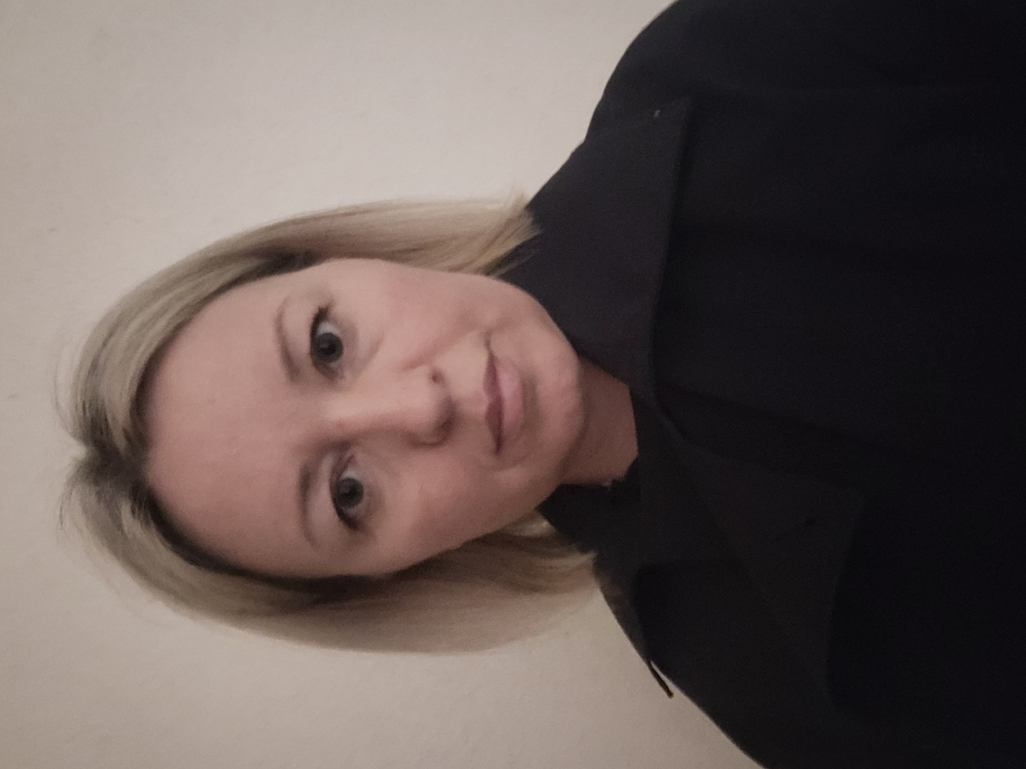 A woman with short blonde hair is posing for a selfie. She is wearing a dark-colored jacket or coat with a collar. The background is a plain, light-colored wall. No information is available regarding her location in Izhevsk, Russia.