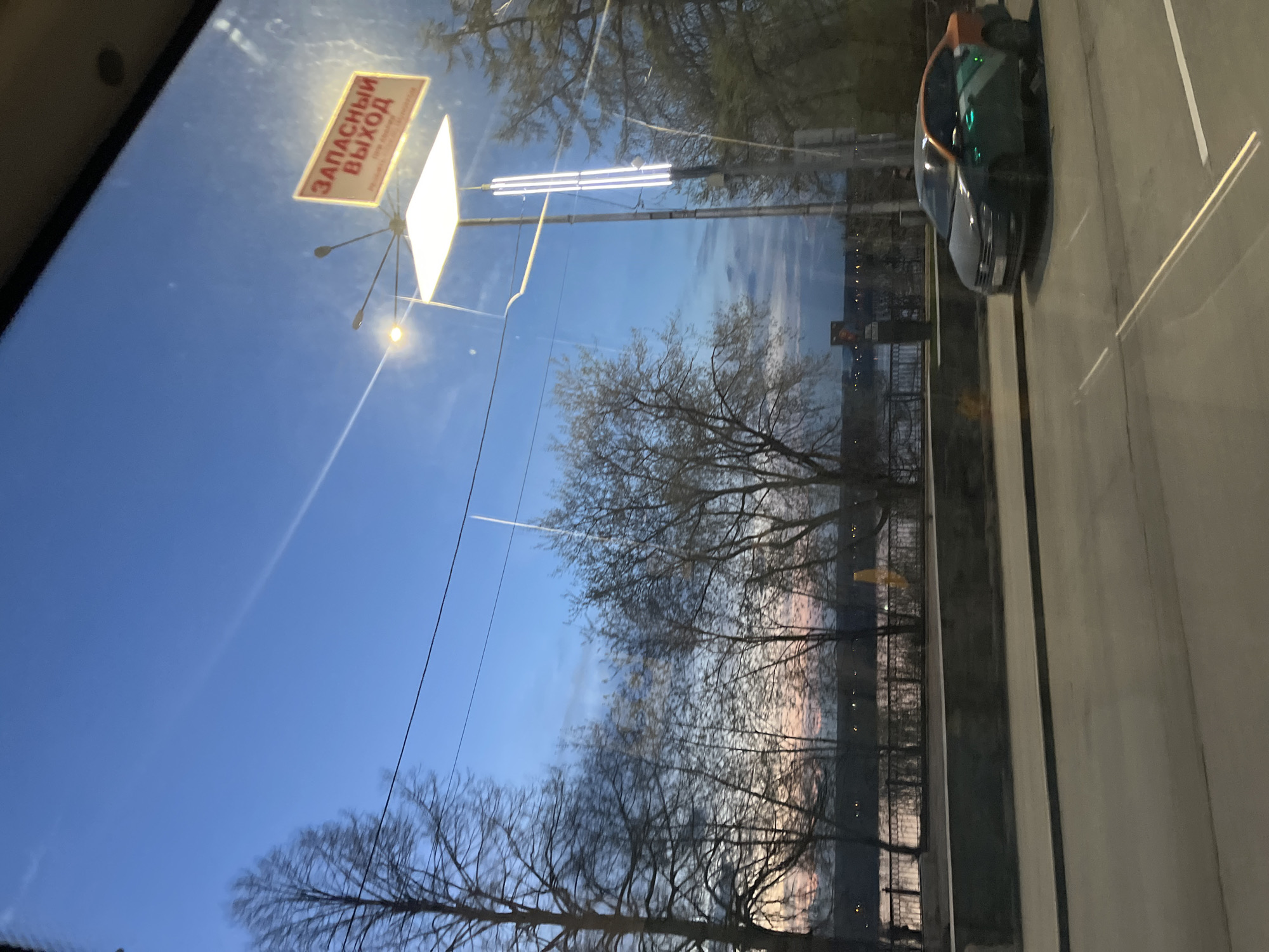 The image depicts a view from inside a vehicle, likely a bus or tram, in what appears to be the city of Perm, Russia. A sign with the Cyrillic inscription "ЗАПАСНЫЙ ВЫХОД" (Emergency Exit) is visible. The sky is a deep blue, transitioning to lighter shades near the horizon, suggestive of either dawn or dusk. Bare trees are silhouetted against the sky and light-colored horizon. A row of lights are observed along the road. An orange-topped vehicle is stopped or parked on the side of the road. A metal fence separates the road from a body of water. The image is shot through a window, indicating the perspective is from inside the vehicle.