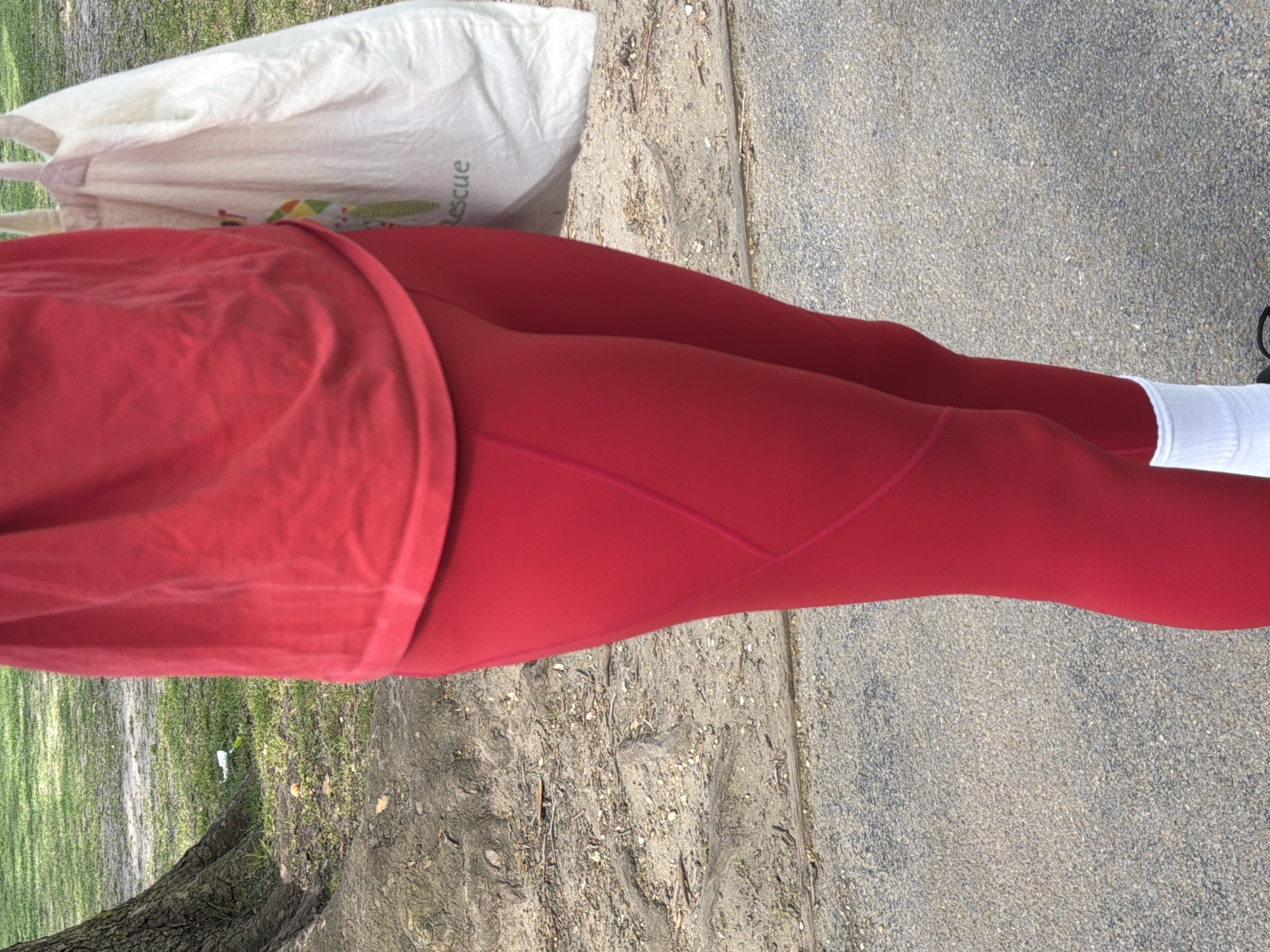 The image shows a person from a slightly elevated perspective, focusing on their lower body as they stand or walk outdoors. The subject is wearing a bright red top and form-fitting red leggings, paired with white socks and partially visible dark-colored shoes on the right foot. The attire suggests athletic or casual wear.
The setting appears to be a park or an outdoor path, characterized by a mix of natural and man-made elements. The ground is composed of a dirt patch with some light green grass on the left side, leading into a textured, grey paved surface resembling asphalt or concrete on the right. The base of a tree trunk and more green grass are visible further to the left, indicating a natural environment.
Behind the person, on the dirt ground, a white canvas bag is discernible. The word "Rescue" is printed vertically in green text on the side of the bag, with some colorful graphics appearing below it. The scene is well-lit by natural sunlight, suggesting it is daytime with clear weather. The overall impression is a casual outdoor activity in a green space within an urban context, consistent with parks found in London, United Kingdom.
