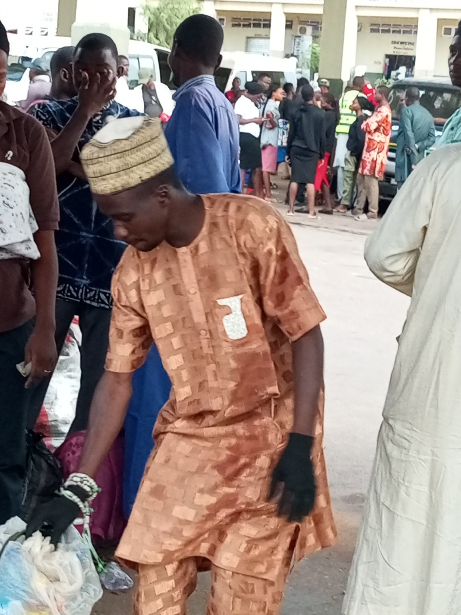 A group of people are gathered outdoors in what appears to be a public area with buildings in the background. One man in the foreground is wearing a traditional bronze-colored outfit with a checkered pattern and a white and gold cap. He is wearing a black glove on his right hand and appears to be reaching into a bag. Another person in a light-colored garment stands to his right. In the background, several individuals are standing and interacting, with vehicles visible. The environment suggests an urban setting. The individuals are of diverse ages and genders, some are dressed in traditional attire, others in more contemporary clothing. The scene is likely in Gombe, Nigeria, given the visual cues and context.