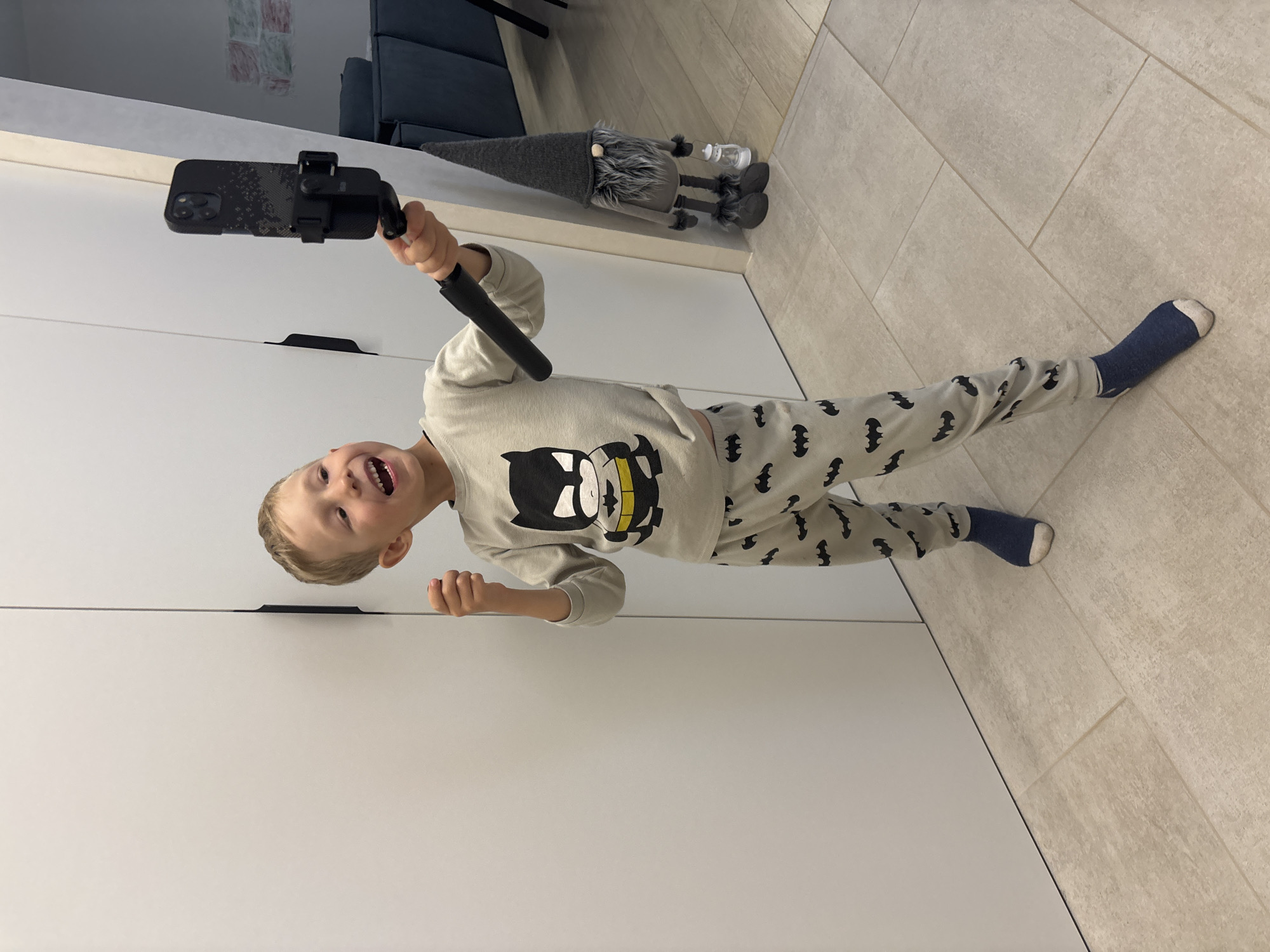 A young boy, likely around 4-6 years old, is captured in a playful moment, holding a smartphone on a selfie stick. He is wearing a light grey long-sleeved shirt with a Batman graphic and matching pants adorned with small black bat patterns. He has on dark blue socks. The boy's face is animated with a wide smile and open mouth, suggesting excitement or laughter. His blond hair is short and tousled.
The scene appears to be indoors, possibly a home. Behind the boy, the environment suggests a modern living space. To his left, large white cabinets or doors are visible, featuring sleek, dark handles. To his right, a tiled floor in neutral tones is seen, with a faint grout pattern. In the background, a grey, soft-textured gnome figurine with a long conical hat and a white lamp is resting on a ledge or piece of furniture. Further in the background, hints of a blue sofa and a wall with abstract artwork suggest a living area.
The lighting is bright, indicating it could be daytime. The boy's energetic pose and expression imply he is actively engaged in taking a selfie or recording a video. The overall mood is joyful and spontaneous. There is no visible text in the image. The location context provided is Chaikovsky, Russia.