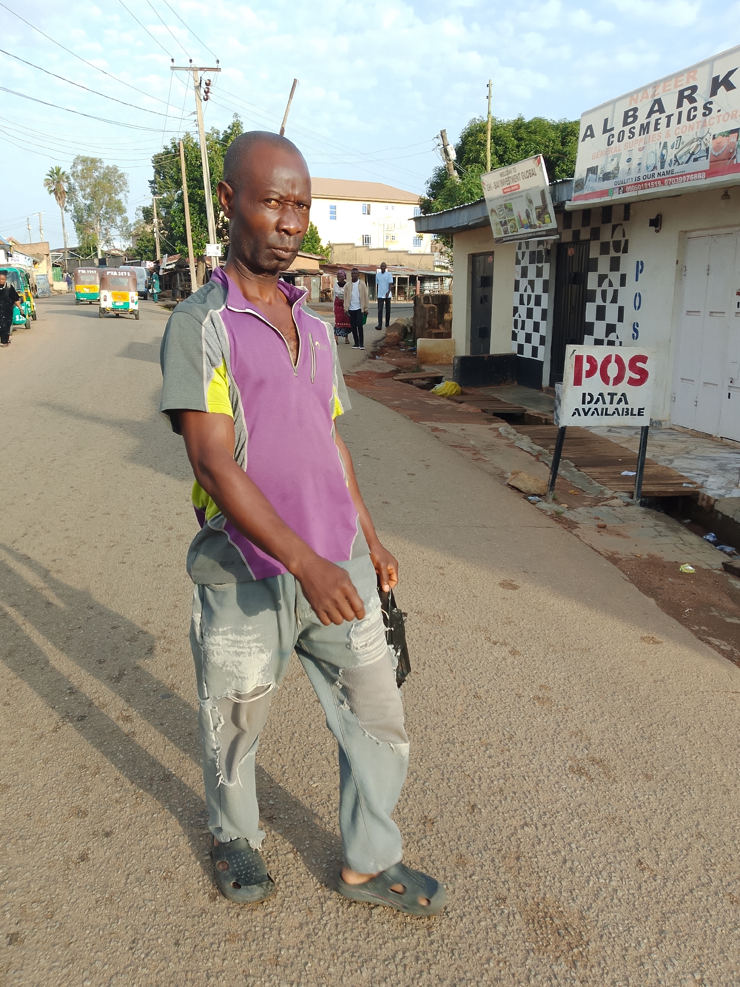 A man in a purple and gray shirt and ripped jeans stands on a paved road, looking at the camera with a stern expression. He is wearing dark clogs on his feet. Behind him, a street lined with buildings and shops is visible. A sign for "NAZEER ALBARK COSMETICS" is prominent on one building. Another sign reads "POS DATA AVAILABLE". In the background, several tricycles, also known as "keke napep," are seen on the road, indicating a busy street scene in the city of Jos, Nigeria. The sky is partly cloudy, suggesting it is daytime. The overall environment appears to be a commercial area in a Nigerian city.