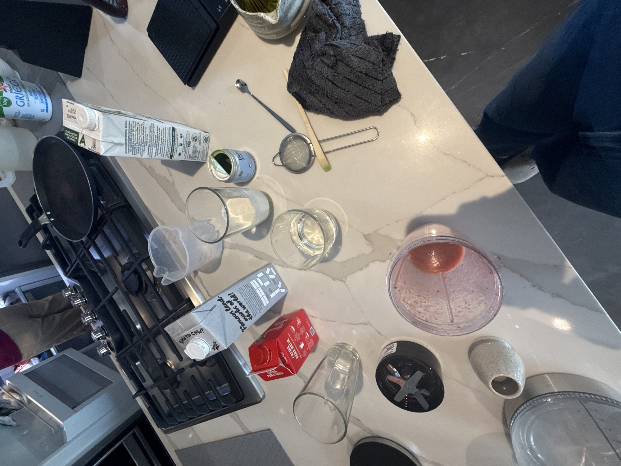 A countertop scene in the city of San Bernardino Tlaxcalancingo, Mexico, shows items likely related to food preparation. A carton of "GRIEGO" yogurt, an organic carton of "SIN AZUCAR" (no sugar) milk, and another carton labeled "CREMA PARA BATIR" (whipping cream) are visible. There are also several glasses, a measuring cup, a tea strainer, and an empty blender. A dark gray towel sits nearby. A stovetop with a black pan sits to the left. A person wearing jeans stands to the right of the countertop.