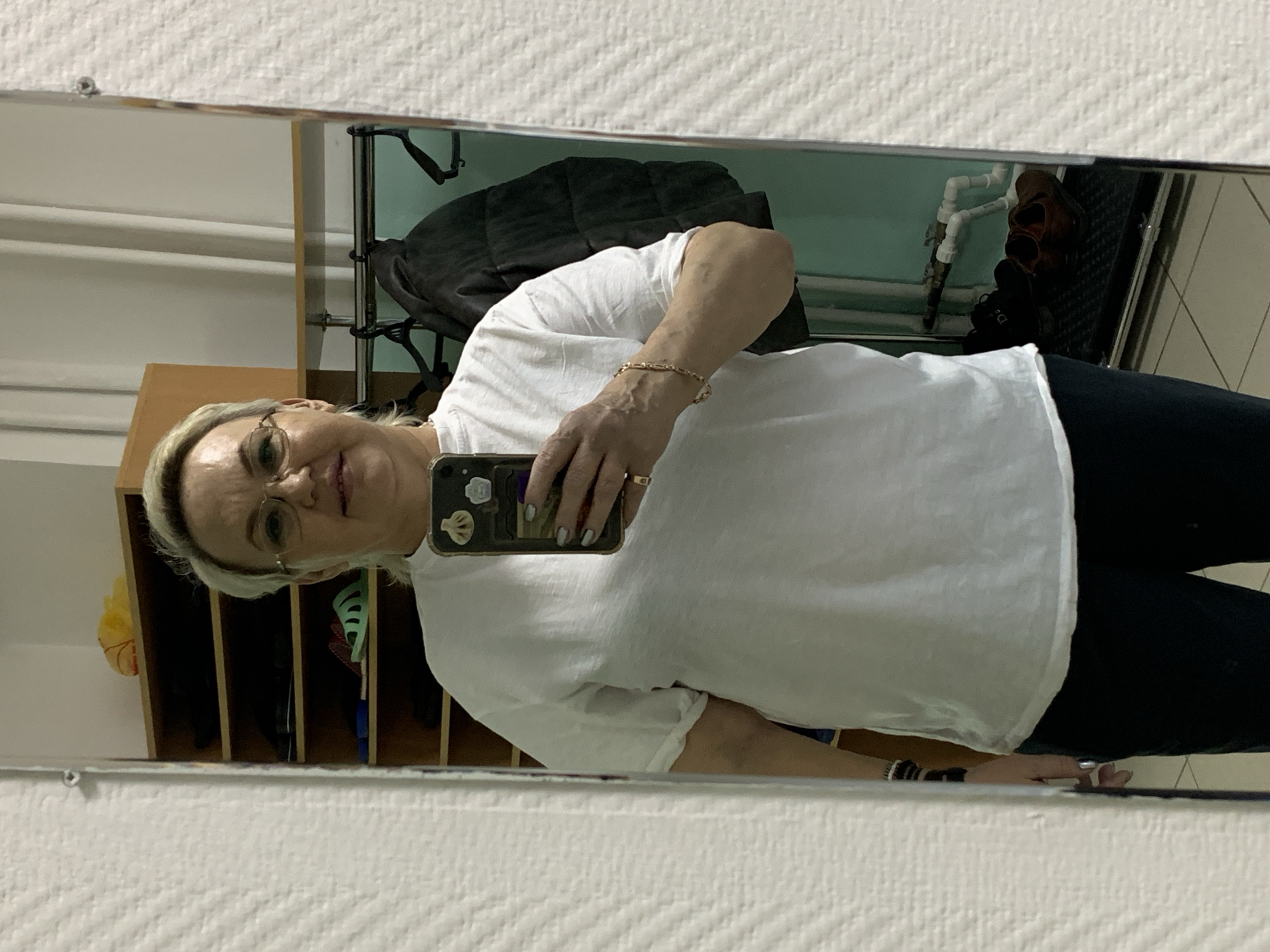 A woman is taking a selfie in a mirror. She is wearing glasses, a white t-shirt, and dark pants. She is holding a black smartphone with a decorated case. The reflection shows a closet with a black coat and a shelf with shoes. The background suggests a public space or hallway. It is not possible to confirm the location is in Chaikovsky, Russia, based on the visual information alone.