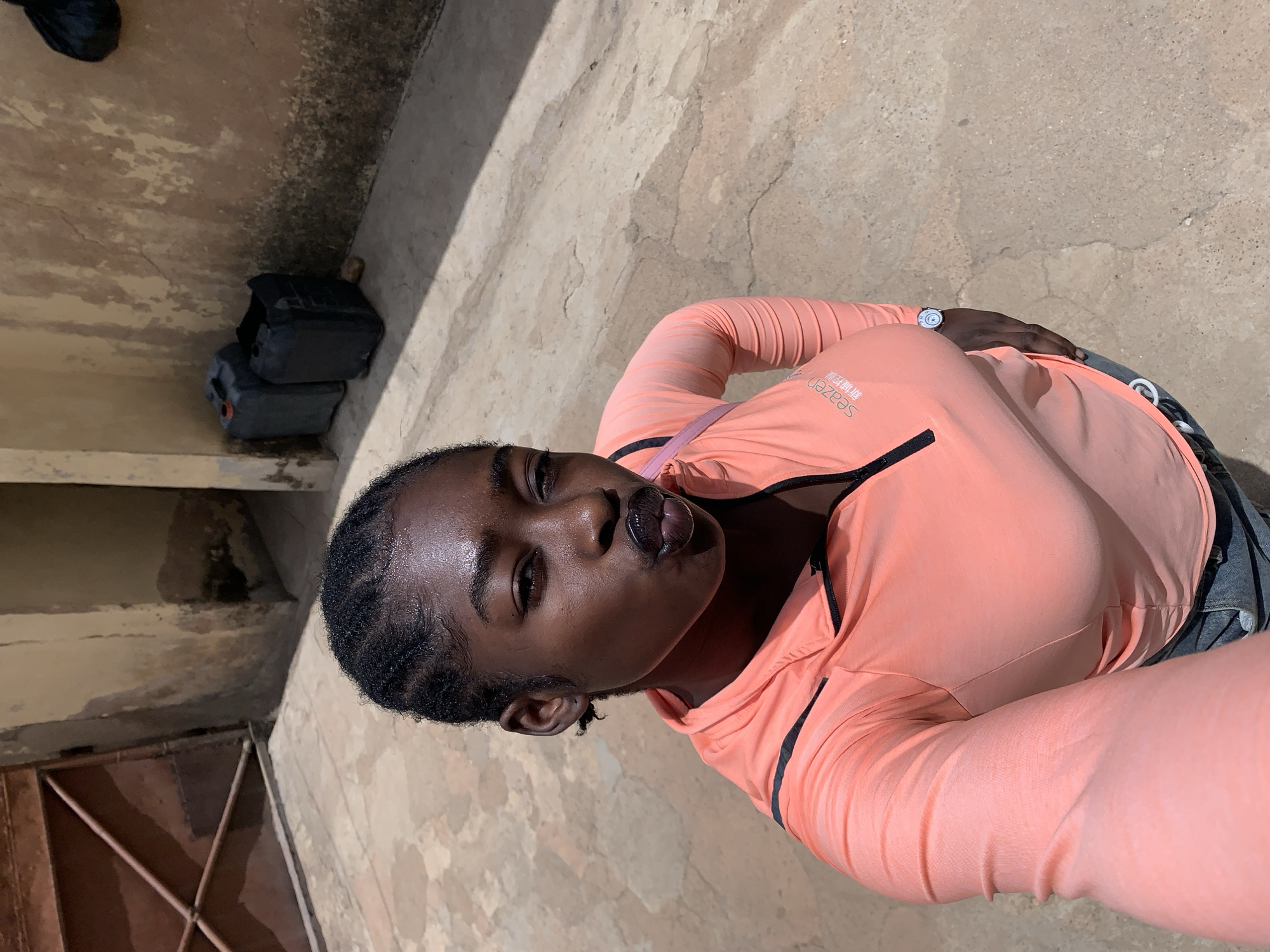 A selfie shot captures a dark-skinned individual with their head tilted to the left, lips pursed. The person is wearing a long-sleeved, coral-colored athletic jacket with black detailing. The background shows a concrete or stone surface, a door with a metallic frame, and dark containers. The location is Rafin Par, Nigeria.