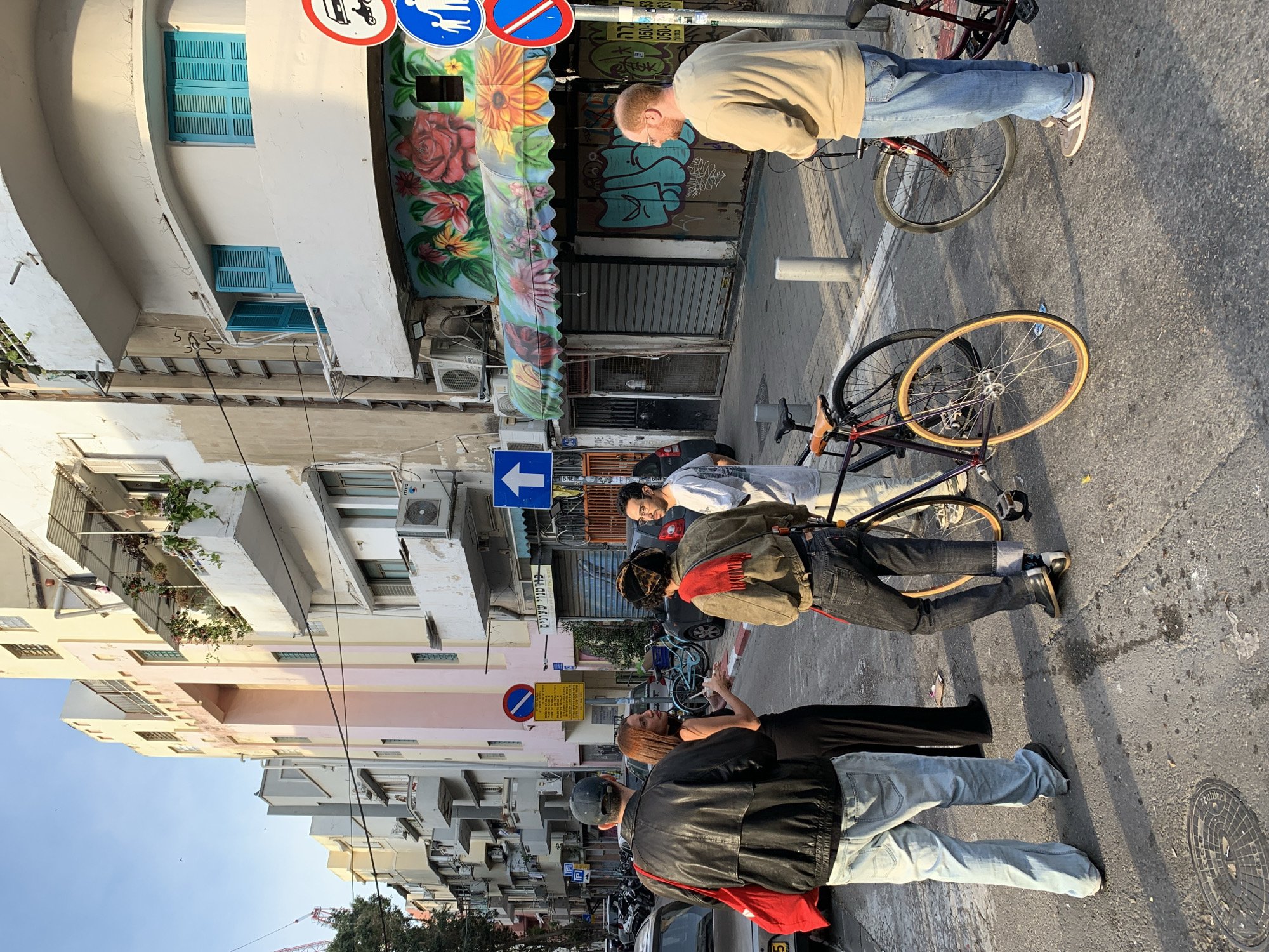 The image shows a street scene in Tel Aviv, Israel. Several people are standing and sitting on the sidewalk and street. One man is on a bicycle, and another bicycle is lying on the ground. Street signs are visible, along with buildings and a shop front with a floral mural. Cars are parked along the side of the street.