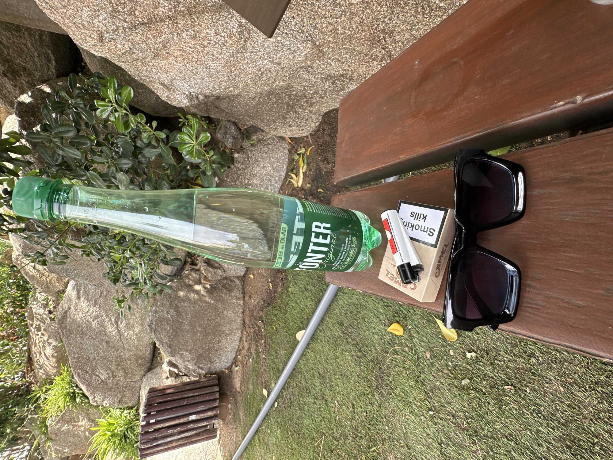 An outdoor scene features a wooden bench with several items placed on it. To the left, a green plastic bottle of Monter Original mineral water lies horizontally, its label partially visible with text including "MONTER Original" and "mineral natural." Next to the bottle, a pack of Camel cigarettes is partially obscured by a pair of black sunglasses. A white lighter with red accents rests on top of the cigarette pack. The bench itself is made of dark wood and appears weathered.
The background consists of large, grey rocks and lush green foliage, suggesting a natural setting. The ground is covered in green artificial turf, with a few scattered yellow leaves. A grey pole runs diagonally across the grass. The scene is illuminated by natural light, indicating it is daytime. The overall impression is a casual, possibly resting spot in a park or outdoor area. The text on the cigarette pack includes a warning: "Smokin kills." The orientation of the image is such that the bottle and sunglasses are oriented from left to right at a downward angle, while the cigarette pack and lighter are positioned slightly above and to the right of the bottle.