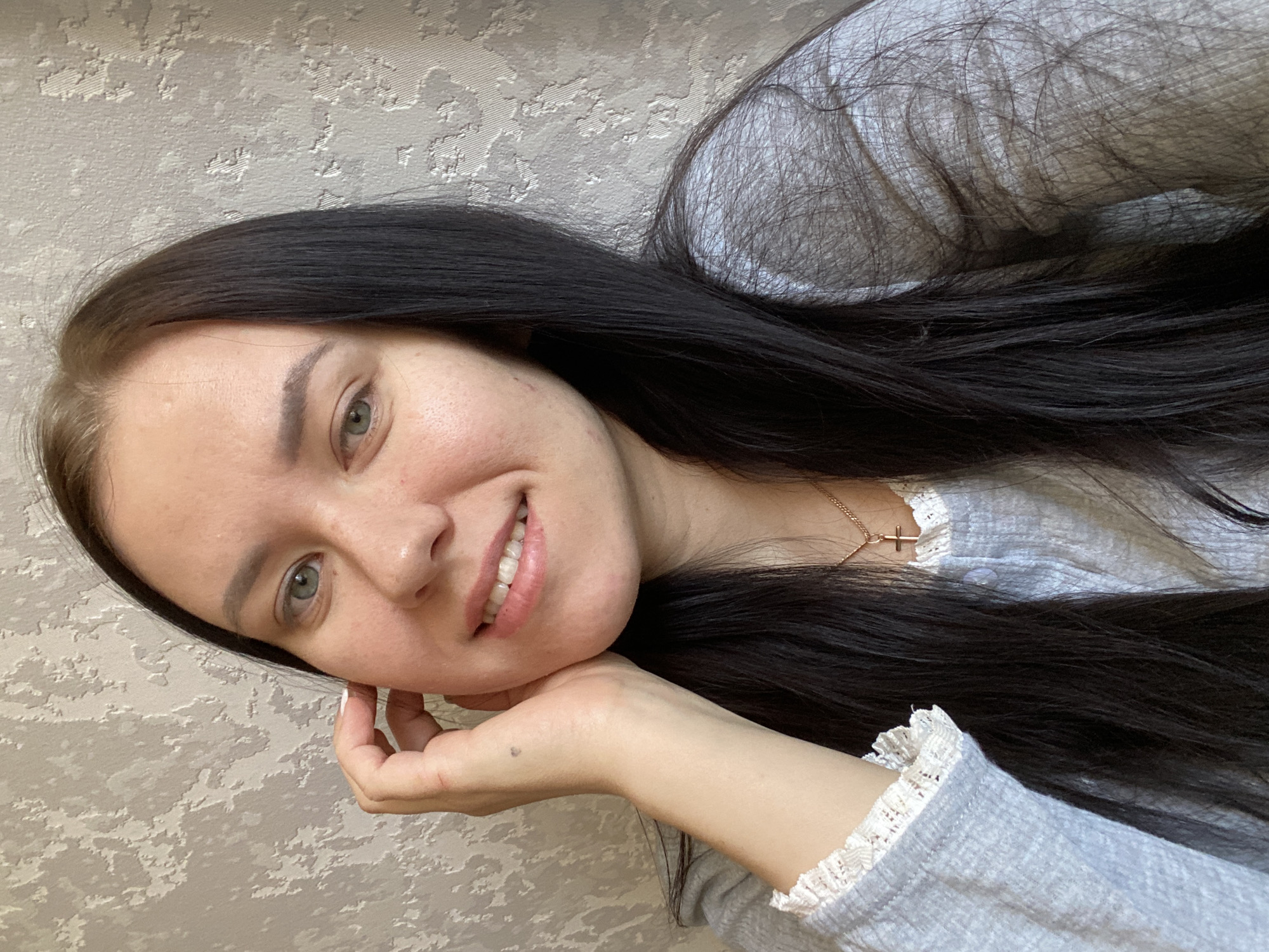 A young woman with long, dark, straight hair and light-colored eyes is captured in a close-up, head-and-shoulders portrait. She is smiling broadly, revealing her teeth, and her head is tilted slightly. Her left hand is gently placed under her chin, with her fingers lightly curled, and a small dark mark is visible on her hand/wrist.
She has a fair complexion, with some minor skin texture visible on her forehead and cheek. Her eyebrows are natural, and her gaze is directed towards the camera.
The woman is wearing a casual light grey long-sleeved top, which features delicate white lace trim along the cuffs and neckline. A thin gold chain with a small cross pendant is visible around her neck.
The background consists of a light-colored, textured wall, likely wallpaper with an abstract, raised pattern, suggesting an indoor setting. The overall lighting is soft and even, indicating daytime. The scene evokes a friendly and relaxed mood. This image was taken in an indoor setting, possibly a home in Chaikovsky, Russia. No visible text is present in the image.