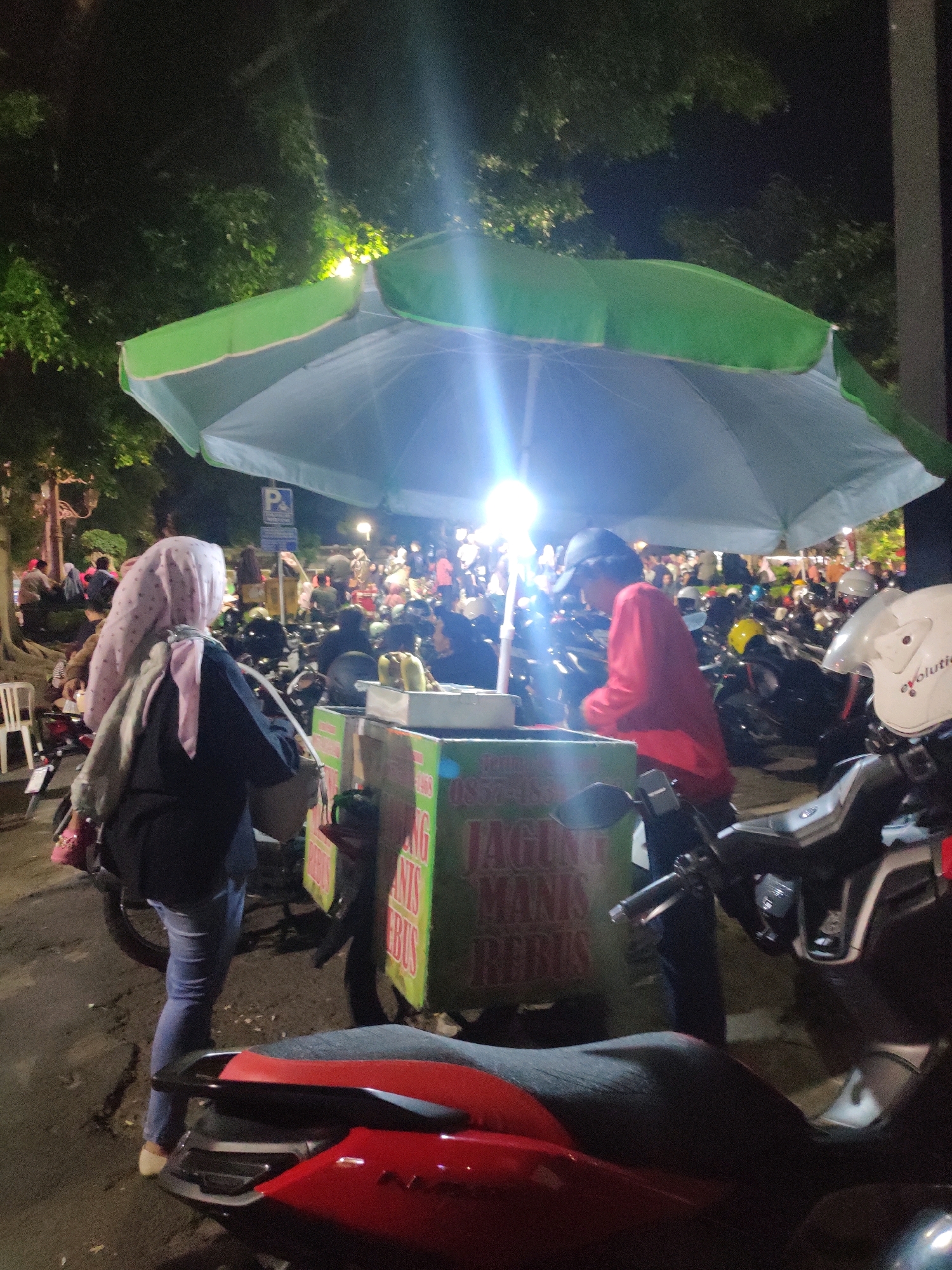 The image depicts a lively night scene at a street food stall in Magetan, Indonesia, likely during a local event or market. The primary focus is on a food cart selling what appears to be corn, indicated by the text on the cart which reads "JAGUNG MANIS REBUS" (sweet boiled corn). The cart is illuminated by bright lights, casting a glow on the surrounding area.
Several people are present, engaging in various activities. In the foreground, a woman wearing a pink headscarf and a dark jacket is seen from behind, walking away from the camera. A person in a red long-sleeved shirt and a black cap is attending to the food cart. Behind them, a crowd of people is visible, many of whom are on motorcycles, suggesting a popular gathering spot. The environment is outdoors, with trees and foliage visible in the background, illuminated by artificial lights. The ground is paved, with some areas appearing to be asphalt. The overall impression is one of a bustling, community-oriented event.