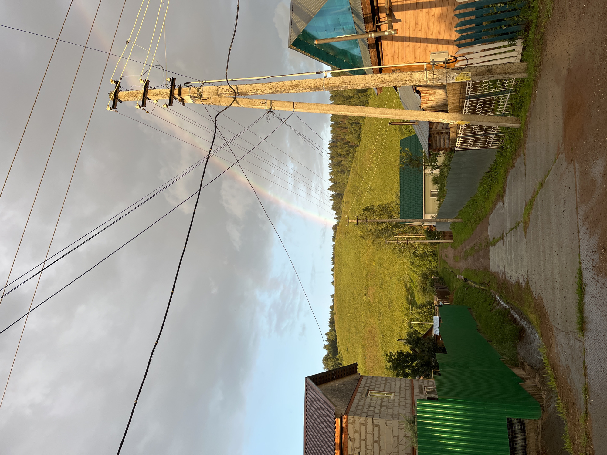 This is a vertical, wide-angle photograph taken in a rural setting in Olkhovka, Russia. The scene depicts a village road with houses and power lines. A subtle rainbow is visible in the overcast sky, suggesting recent rain. The time of day appears to be late afternoon or early evening, as indicated by the warm lighting on the wooden house on the right. The weather is likely humid and possibly still raining, with the road showing signs of wetness.
The foreground features a section of a road made of concrete slabs and dirt, with green grass growing in patches. Several power lines stretch across the frame, supported by wooden utility poles. On the right side of the road, a wooden house with a blue corrugated metal roof and wooden siding is partially visible. Further down the road, more houses with green metal roofs and a white metal fence can be seen.
The background is dominated by a sloping green field that transitions into a dense line of trees. A light rainbow arcs across the sky, partially obscured by clouds. The overall mood is peaceful and quiet, characteristic of a rural landscape after a rain shower. There is no visible text in the image.