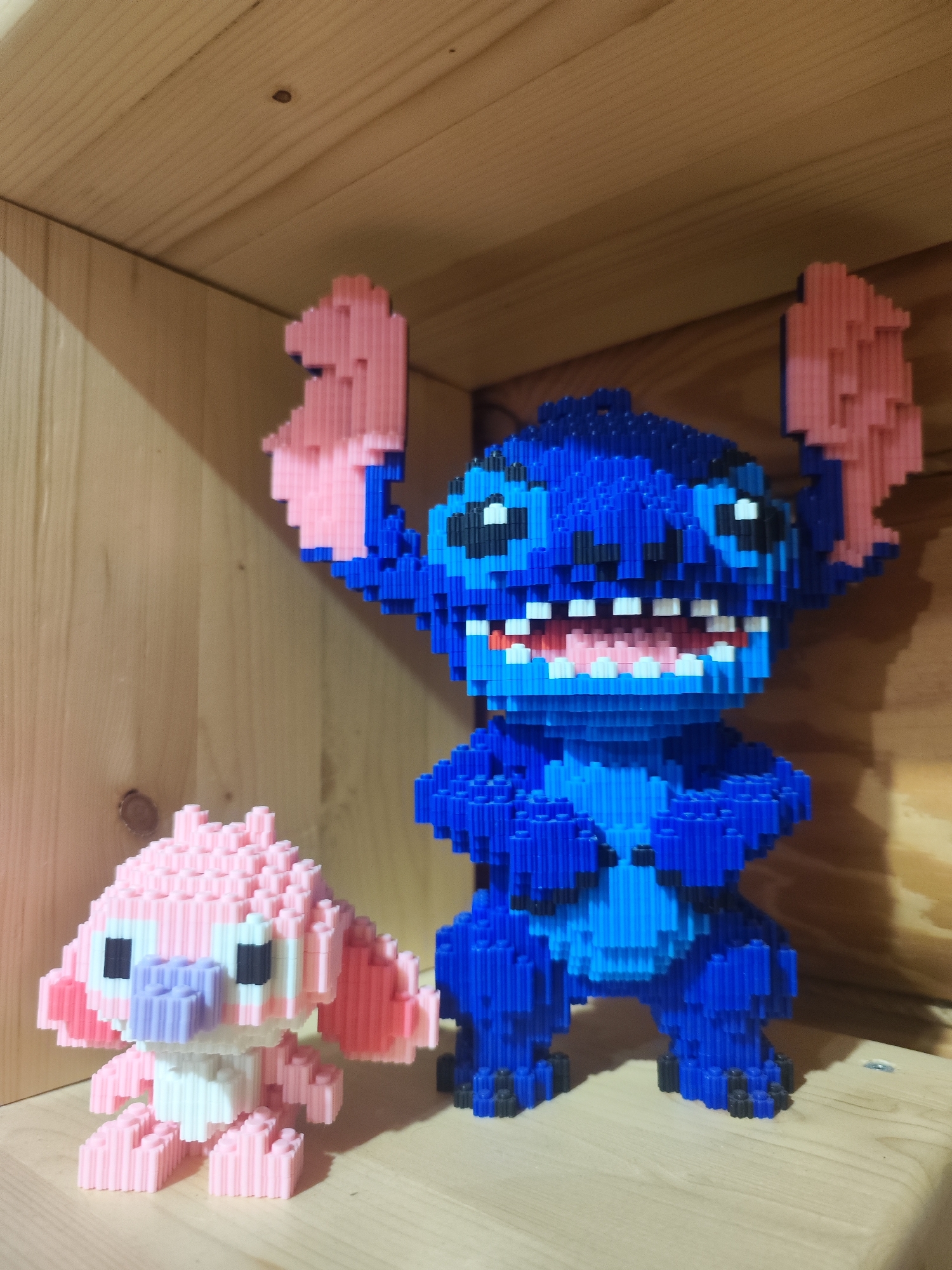 The image shows two block-built figurines, likely toys or collectibles. One is blue and appears to be Stitch from Disney's "Lilo & Stitch". The other is pink and also appears to be a character from the same franchise, possibly Angel. The figurines are standing on a light-colored wooden surface, within what appears to be a wooden shelf or enclosure. Based on the information provided, the location is in Chaikovsky, Russia.