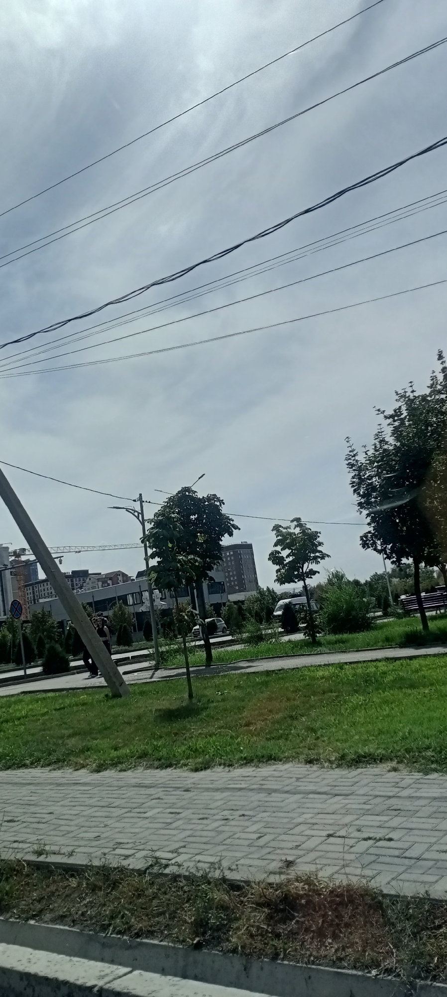 The image depicts a view of a street scene in Bishkek City, Kyrgyzstan, under a cloudy sky. In the foreground, a paved walkway with a grassy area beside it. A concrete pole is prominently visible on the left side of the frame, leaning slightly.
Further into the scene, a street runs parallel to the walkway, with a few cars visible. Trees and bushes are planted along the street and sidewalks, providing greenery. In the background, several multi-story buildings, including what appear to be residential or office complexes, are present, with construction cranes visible near some of them, indicating ongoing development.
The presence of overhead electrical wires crisscrossing the sky is a notable feature of the urban landscape. A few individuals are walking on the sidewalk. The overall impression is that of a typical day in a developing urban area.