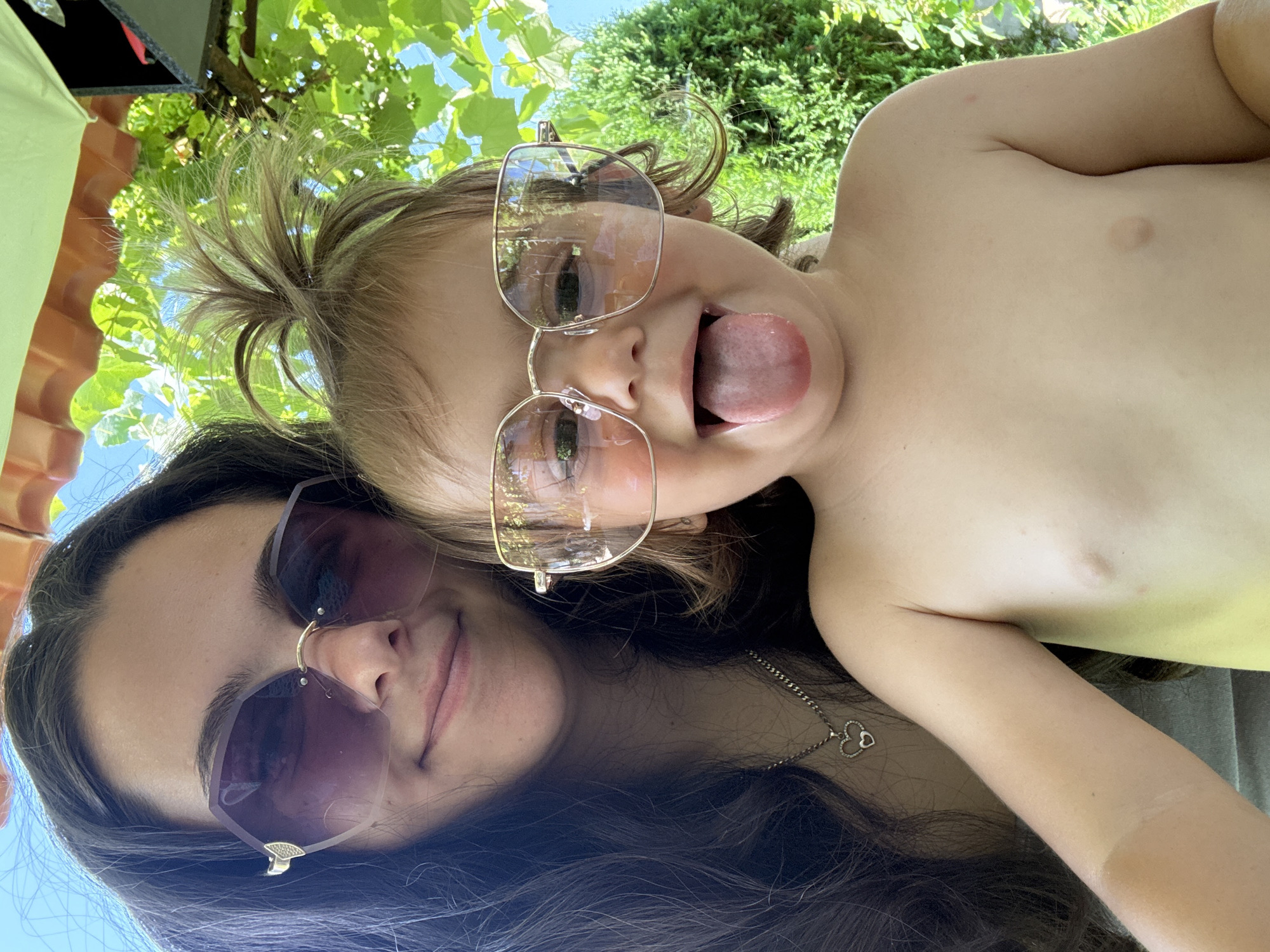 In Oreshak, Bulgaria, two individuals, an adult female and a young child, are posing for a selfie outdoors. The adult female, with long dark hair and wearing large, hexagonal purple sunglasses, is smiling. The child, in the foreground, is also wearing large, square-shaped gold-rimmed sunglasses and is sticking out their tongue. The child is shirtless. The background features lush green foliage and a bright blue sky, suggesting a sunny day. The scene appears to be set in a garden or outdoor seating area, possibly under a canopy indicated by the presence of a roof-like structure.