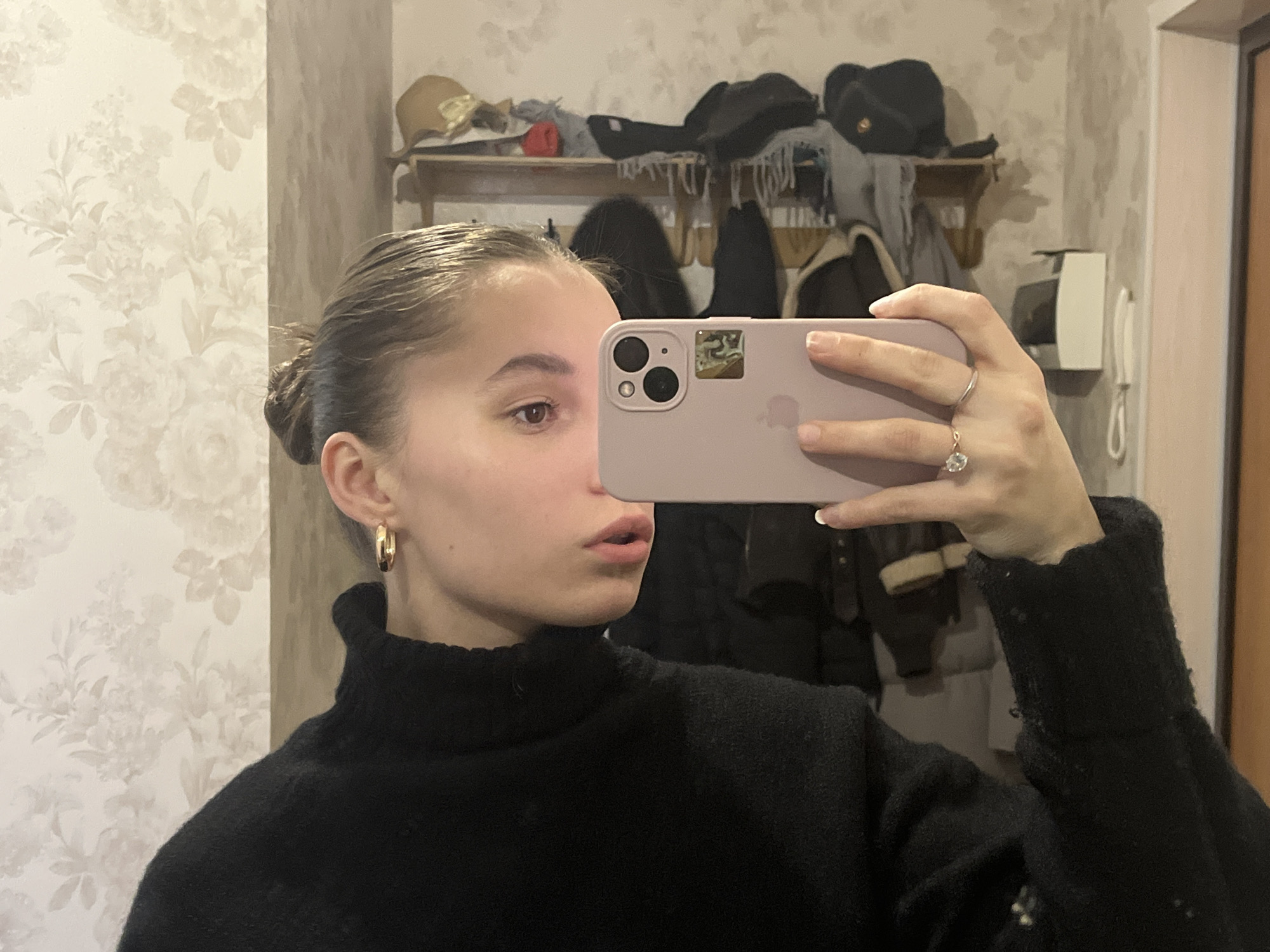 This mirror selfie captures a young woman in an indoor setting, likely a hallway or entryway, in Chaikovsky, Russia. The central subject is a woman, appearing to be in her late teens or early twenties, with light skin, brown eyes, and dark hair pulled back into a bun. She is dressed in a black turtleneck sweater and wears gold-colored hoop earrings. On her left hand, she has a ring with a prominent clear stone on her ring finger and a plain band on her middle finger. Her nails feature a subtle, light polish.
She holds a light purple smartphone, equipped with a triple camera system, in her right hand, using it to take the photograph. The phone partially covers the lower-left side of her face. Her lips are slightly parted, and her gaze is directed towards her reflection or the phone, conveying a poised or thoughtful expression.
The background, visible through the mirror, offers glimpses of the room's decor. The wall to the left is covered with light-colored floral wallpaper, showcasing cream or beige flower patterns. To the right, a wooden shelf holds various items, including a beige hat, a black fur-like hat, and multiple scarves or pieces of outerwear, indicative of a coat rack or storage area. Further right, a white wall-mounted intercom or telephone unit is visible against a plain, light-colored wall. The indoor lighting suggests no specific time of day, but the warm clothing implies cooler weather. There is no visible text within the image.