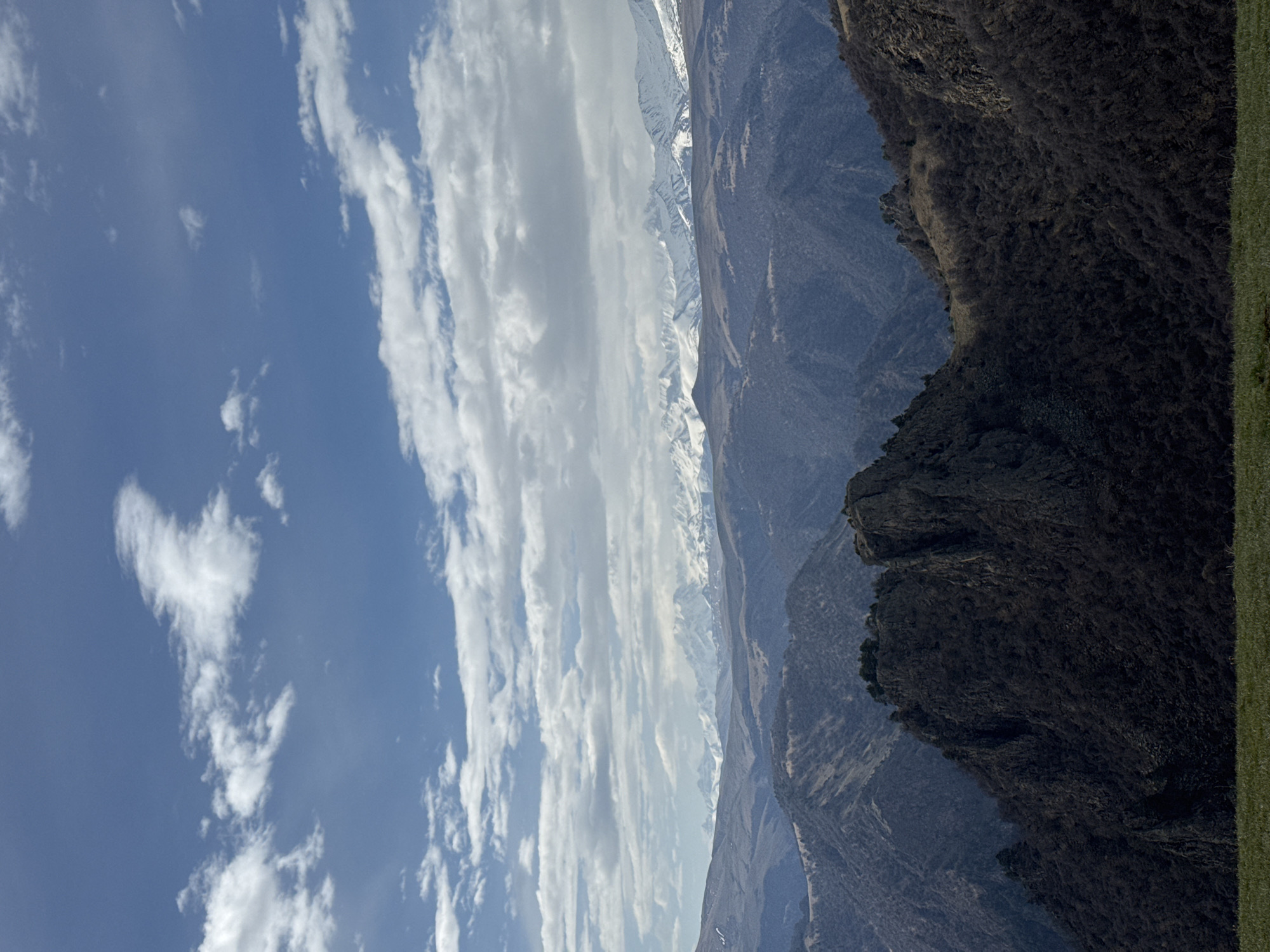 The image depicts a landscape with mountains and sky. The sky is blue with white clouds. A range of snow-capped mountains is visible in the distance. The foreground consists of rocky terrain with sparse vegetation. The image is taken from a high vantage point. Location: Unknown, Russia.