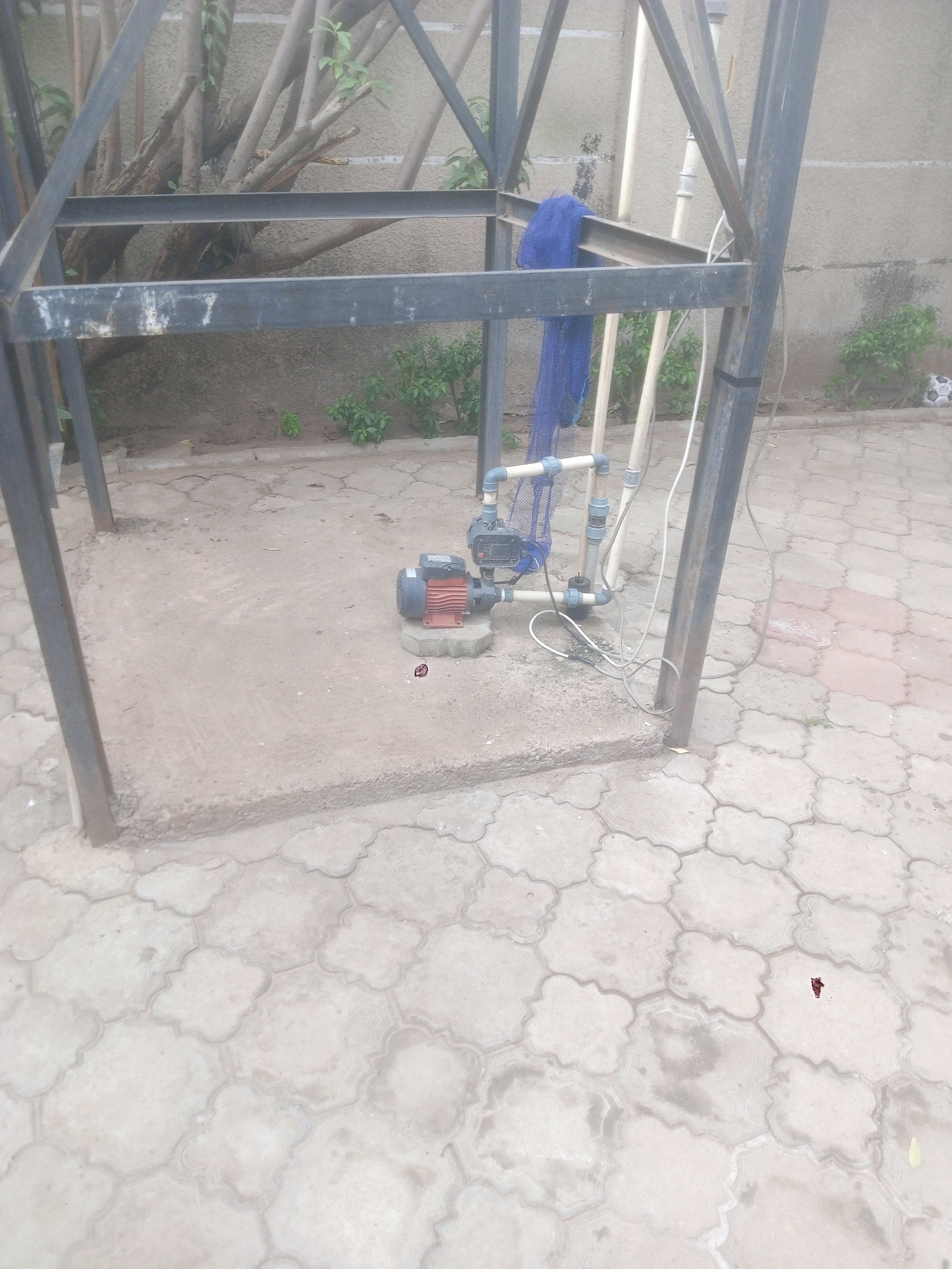 The image shows a water pump installation within a metal frame structure, likely for a domestic water supply system. The location appears to be an outdoor area within Kubwa, Nigeria, as indicated by the style of the paving stones and general environment. The pump is positioned on a concrete base, with PVC pipes connected to it. A blue net is draped over a section of the metal frame. Electrical cables are visible, extending from the pump. The surrounding area is paved with interlocking concrete pavers. Plant growth is visible in the background, along a wall. The overall environment suggests a residential setting. There are no people visible in the image.