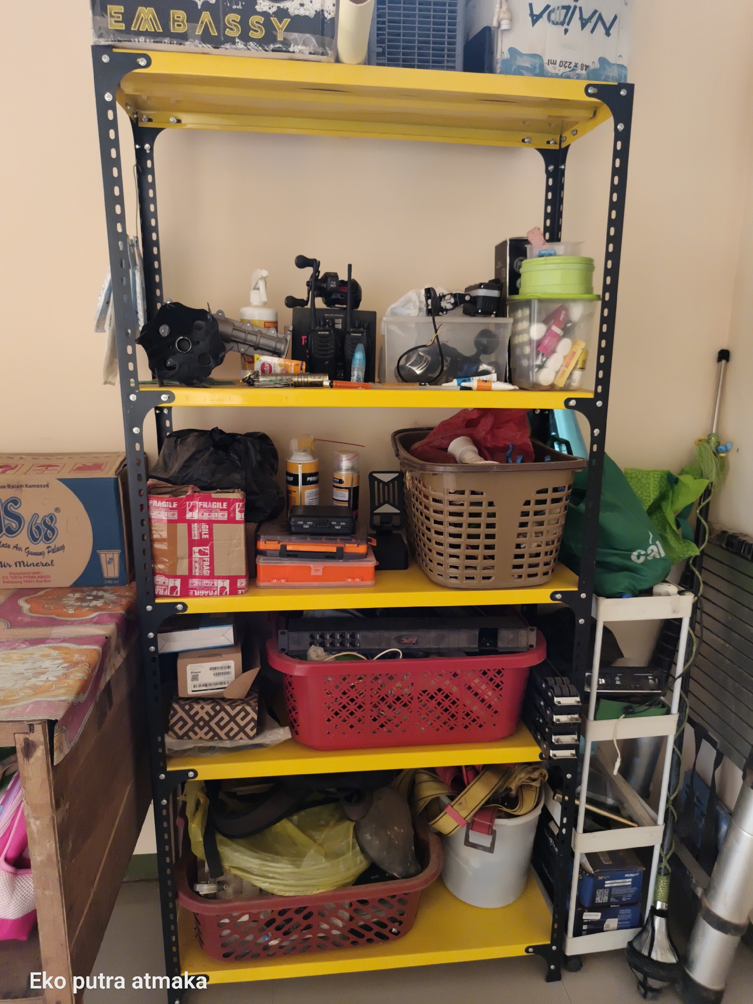 The image shows a yellow metal shelving unit filled with various items. On the top shelf, there are boxes and a bottle of water. The second shelf contains miscellaneous objects including a black fan, a spray bottle, and two walkie-talkies. The third shelf holds more items like a black garbage bag, a "FRAGILE" taped box, two spray cans, and a small black case on top of an orange plastic box. The fourth shelf features a large brown basket filled with red fabric and a white plastic bucket containing yellow straps. The bottom shelf has a red plastic basket and a white bucket with items inside. To the left of the shelving unit is a portion of a table with a patterned cloth. To the right, a mop leans against the wall, and a green bag is visible. The shelving unit is positioned against a light-colored wall. The text "Eko putra atmaka" is visible at the bottom left of the image, likely a watermark or caption. The scene appears to be an indoor storage area in Pelanjau Jaya, Indonesia, indicated by the text and context, possibly a garage or utility room.