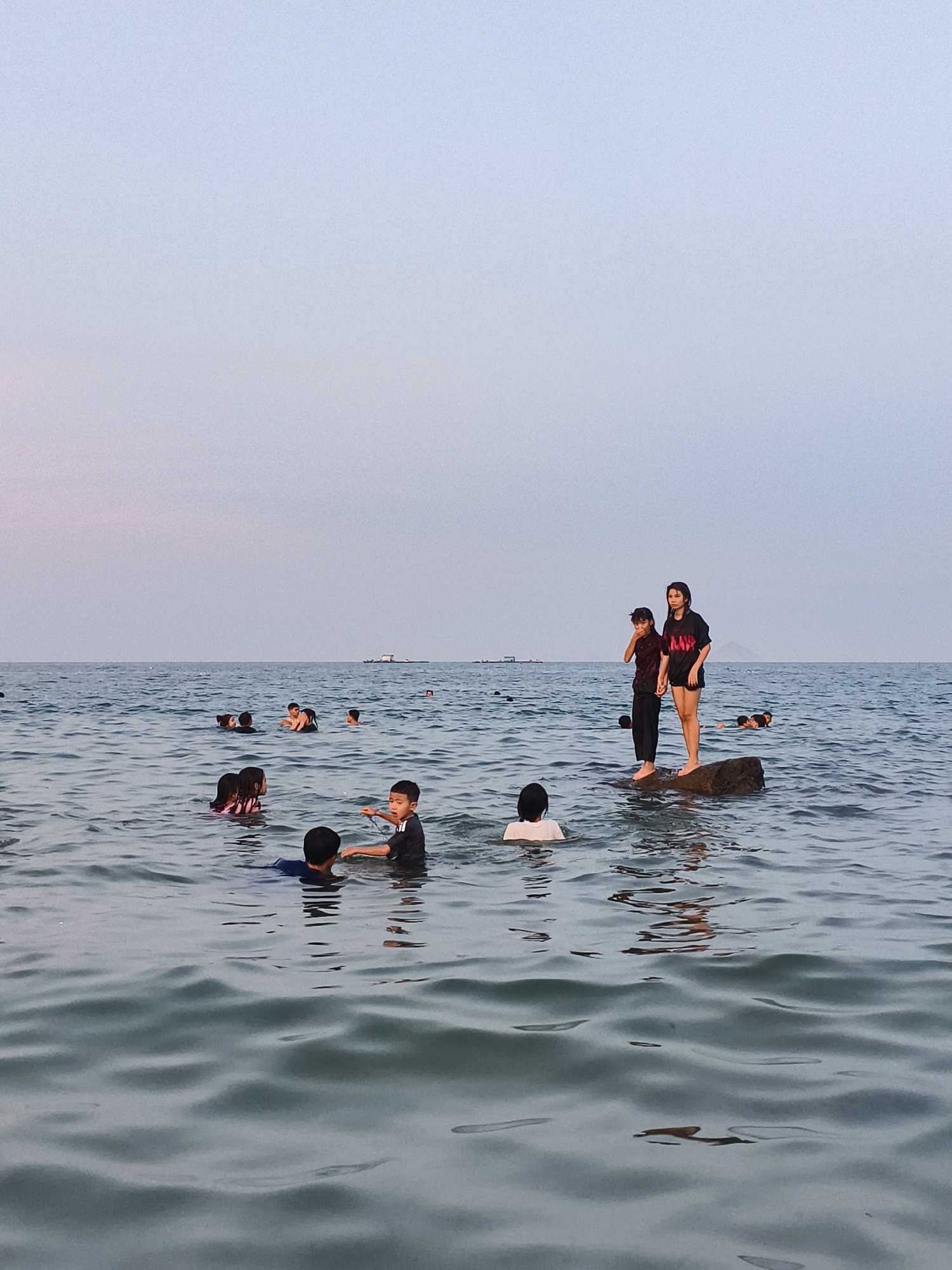 The image shows a group of people, mostly children, swimming in the sea. The sky is overcast with a pale blue and pink gradient. The water is choppy and reflects the sky. In the foreground, two children are visible in the water, one of whom is pointing. In the middle ground, a group of people are swimming, and further back, two individuals are standing on a rock. In the distance, a barge is visible on the horizon. The scene suggests a public beach with recreational activities. Based on the prompt, this scene is located in the city of Vĩnh Lương, Vietnam.