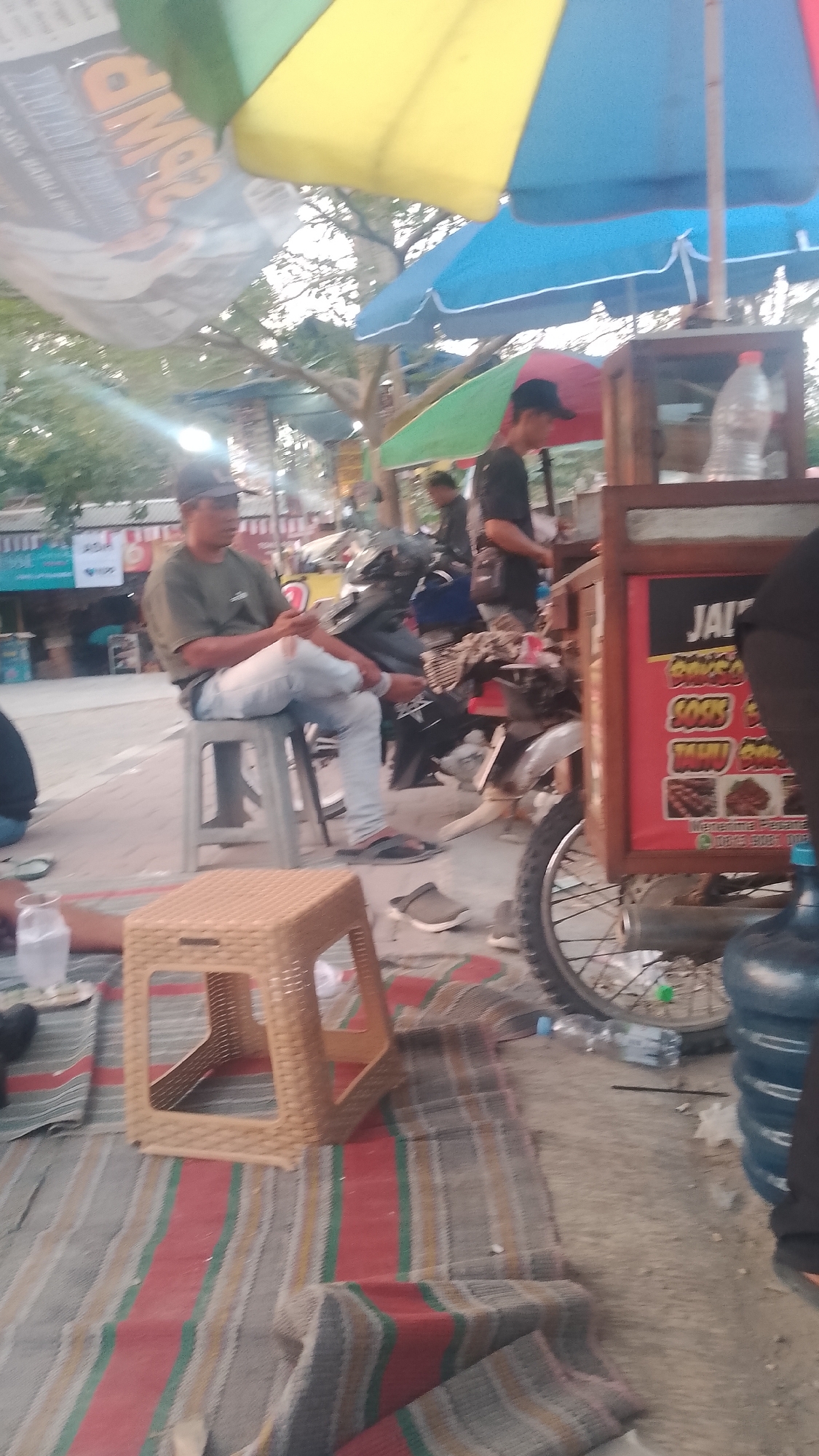 In Karanglangu, Indonesia, a man wearing a green t-shirt and light blue jeans sits on a gray plastic stool. He is looking down at his phone, which he holds in his left hand. His right leg is crossed over his left leg, and he is wearing black flip-flops. In front of him, on a striped mat, is a tan plastic stool. Behind the man, a food cart with a wooden frame and a red sign is visible. The sign has text and images on it, and there is a bottle of water in the cart. A large blue water jug sits on the ground next to the cart. Several umbrellas, including yellow, blue, and green ones, are set up over the area. In the background, there are trees and buildings, suggesting an outdoor market or roadside eatery.