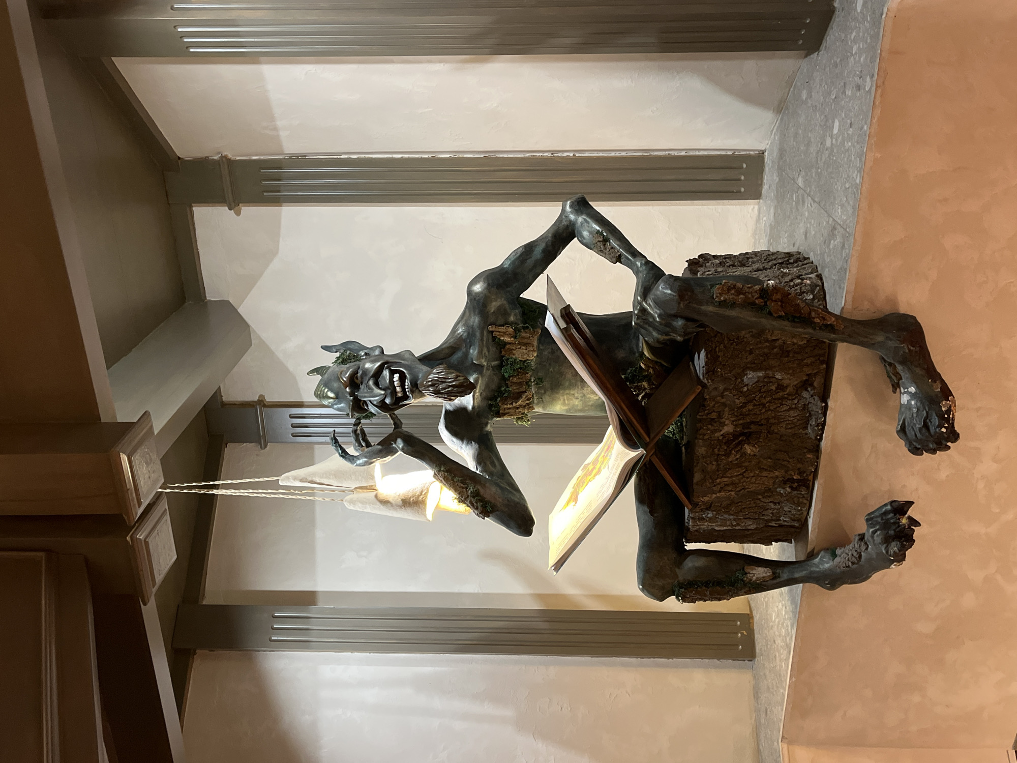A bronze sculpture of a gnome-like figure reading a book sits atop a wooden pedestal in an indoor location. The figure is twisted and animated, with its face bearing a grotesque expression. The sculpture is illuminated by a light source directly behind it. The background features neutral-toned walls with linear architectural detailing and trim. The sculpture is located in Kazan, Russia.
