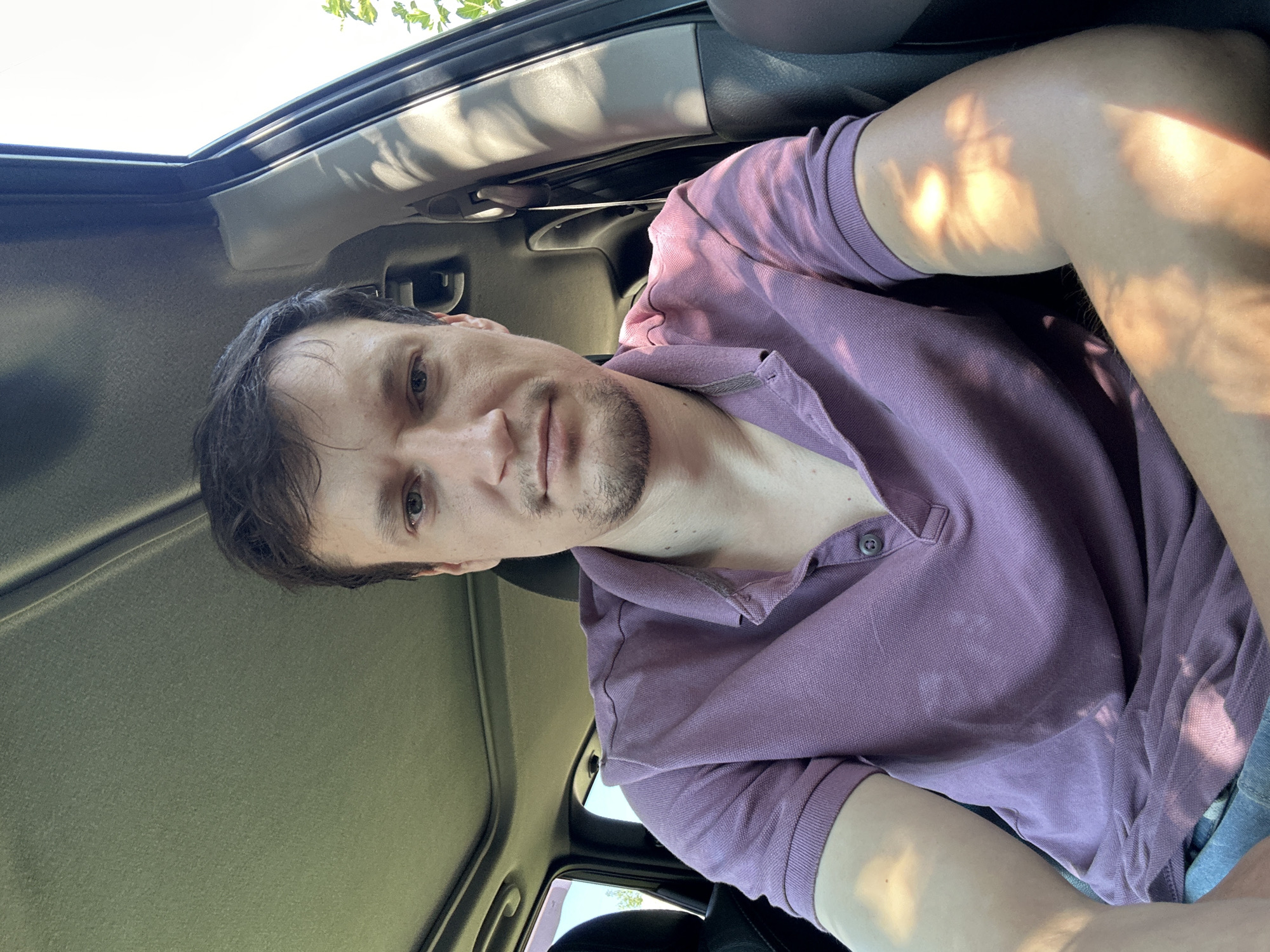 The image shows a man inside a car, possibly in Shatava, Ukraine. He is looking directly at the camera, with a neutral expression. The man is wearing a purple polo shirt. Sunlight streams through the car windows, casting shadows on his face and arm. The interior of the car is visible, including the headliner and part of the dashboard. The scene appears to be during the daytime, with trees and sky visible through the car windows.