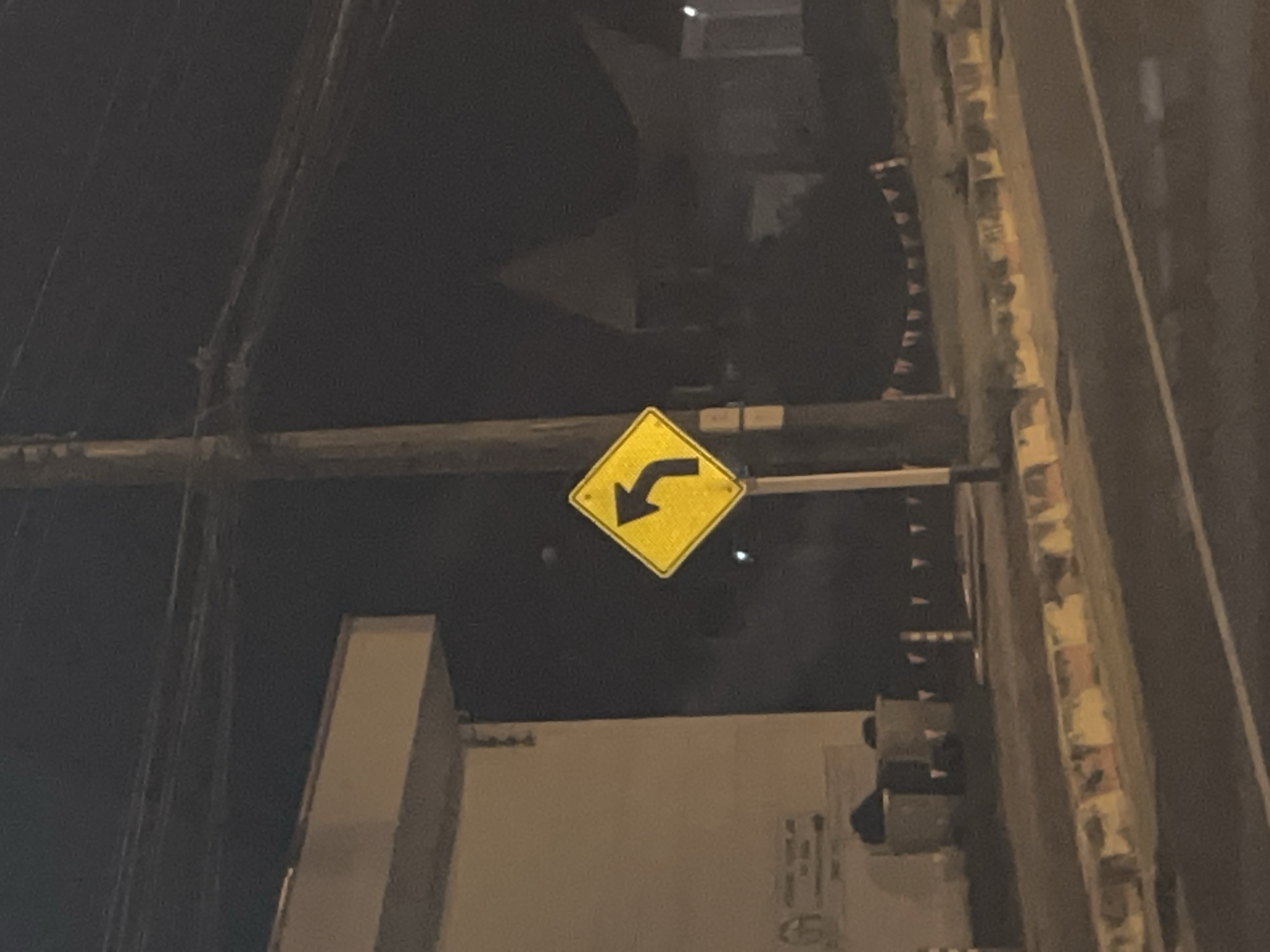 In Chalong, Thailand, at night, a yellow diamond-shaped traffic sign with a black arrow pointing left is mounted on a utility pole. The sign indicates a left curve ahead. To the left of the sign is a portion of a light-colored building. In the background, silhouetted buildings and trees are visible against the dark night sky, with some stringed flags draped across the scene. The foreground shows a road with a curb and sidewalk. The image is slightly blurry due to the low light conditions and possibly motion.
