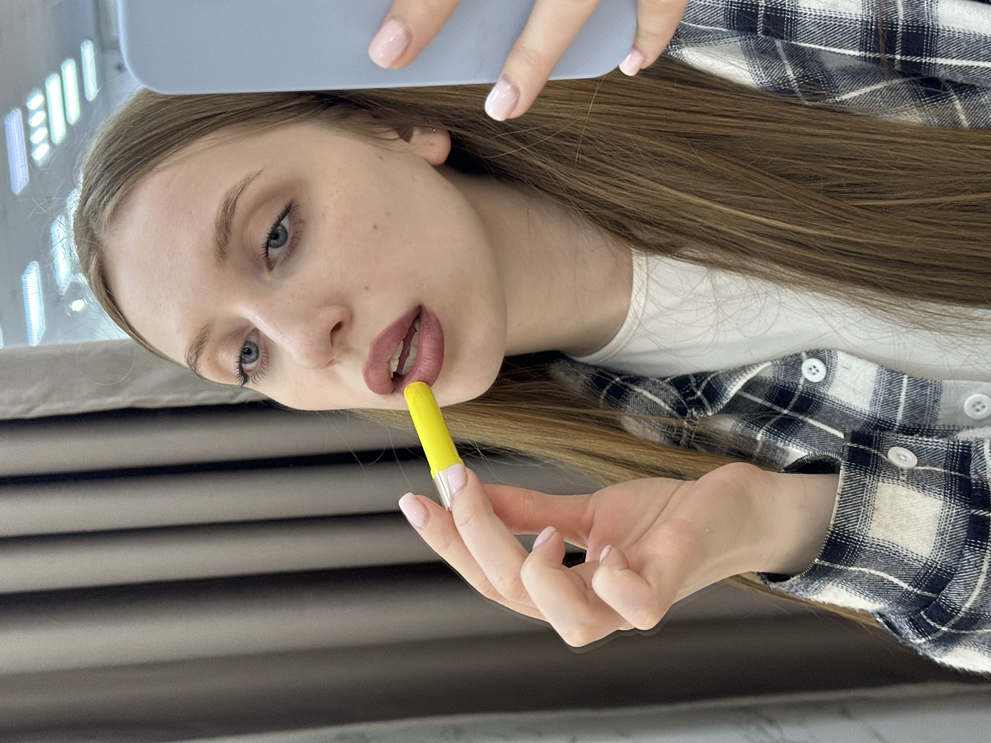 A young woman is depicted in a close-up, chest-up view, actively applying lipstick. Her long, light brown hair falls over her shoulders, framing a face with blue eyes and fair skin exhibiting minimal freckling. Her right hand, with fingernails coated in light pink polish, holds a bright yellow lipstick tube featuring a silver band, applying a reddish-brown shade to her slightly parted upper lip. A light blue smartphone is partially visible on the right side of the frame, held by her left hand, which also displays light pink polished nails. She is dressed in a white crew-neck shirt beneath an unbuttoned black and white plaid flannel shirt with white buttons. The background consists of dark gray, vertically oriented curtains. Reflections of rectangular overhead light fixtures are visible in the upper right quadrant of the image. The specific location within Chaikovsky, Russia, is not discernible from the visual evidence provided.