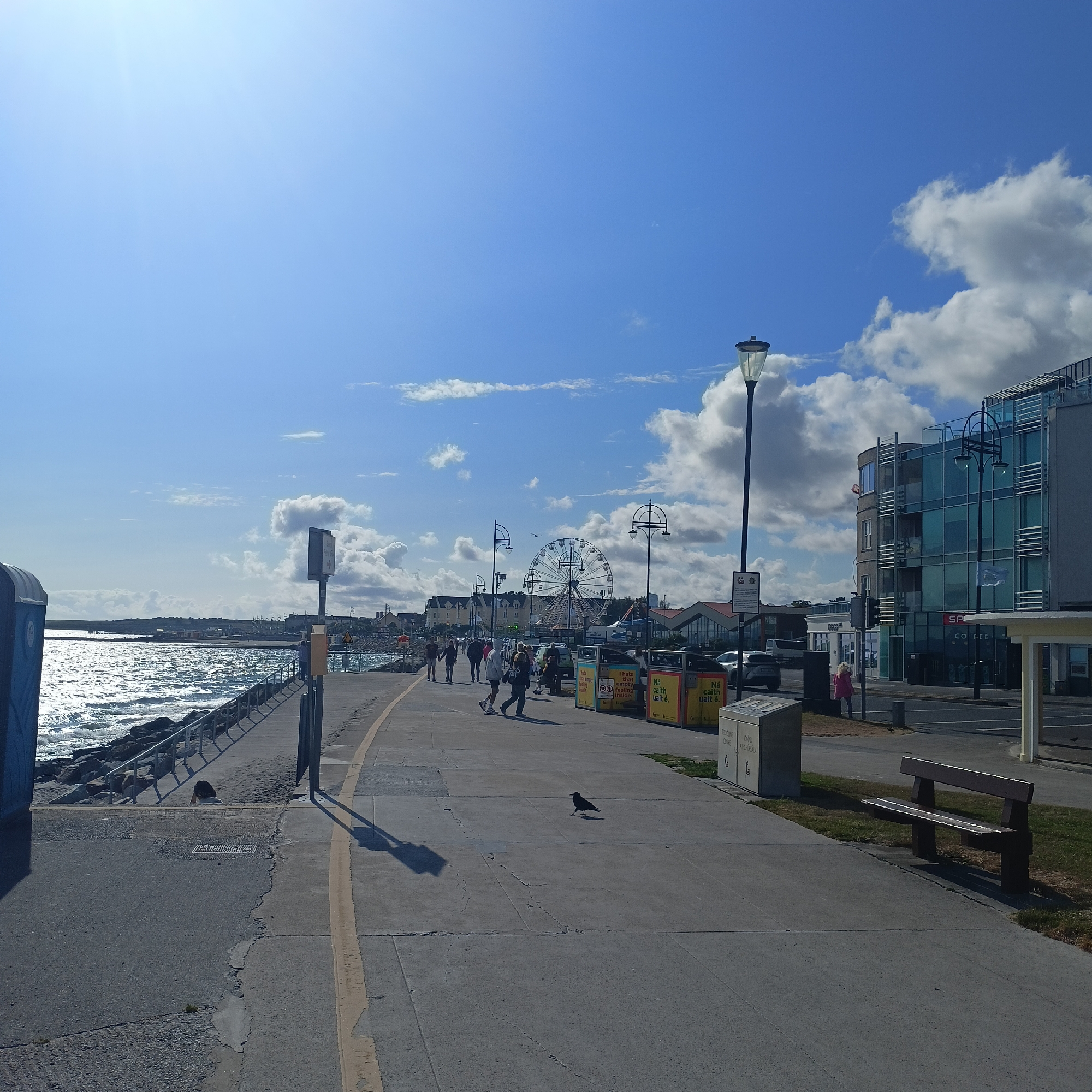 The image depicts a scenic view of the Salthill Promenade in Galway, Ireland, on a bright, sunny day. The promenade runs along the coast, with the Atlantic Ocean to the left, its surface shimmering under the sun. A rocky breakwater lines the edge of the promenade, separating it from the water.
Several people are walking along the promenade, enjoying the weather and the views. Some are dressed casually, while others appear to be tourists. In the distance, a Ferris wheel is visible, a common attraction in seaside towns. Buildings line the right side of the promenade, including a modern glass-fronted building that appears to house shops and cafes. A lamppost stands prominently in the foreground, casting a long shadow across the concrete pathway.
There are also various functional objects present, such as bins, benches, and a portable toilet. A crow is seen standing on the pavement, adding a touch of nature to the urban landscape. The sky is a vibrant blue, dotted with fluffy white clouds. The overall atmosphere is one of a pleasant day out, with people enjoying leisure activities in a beautiful coastal setting.