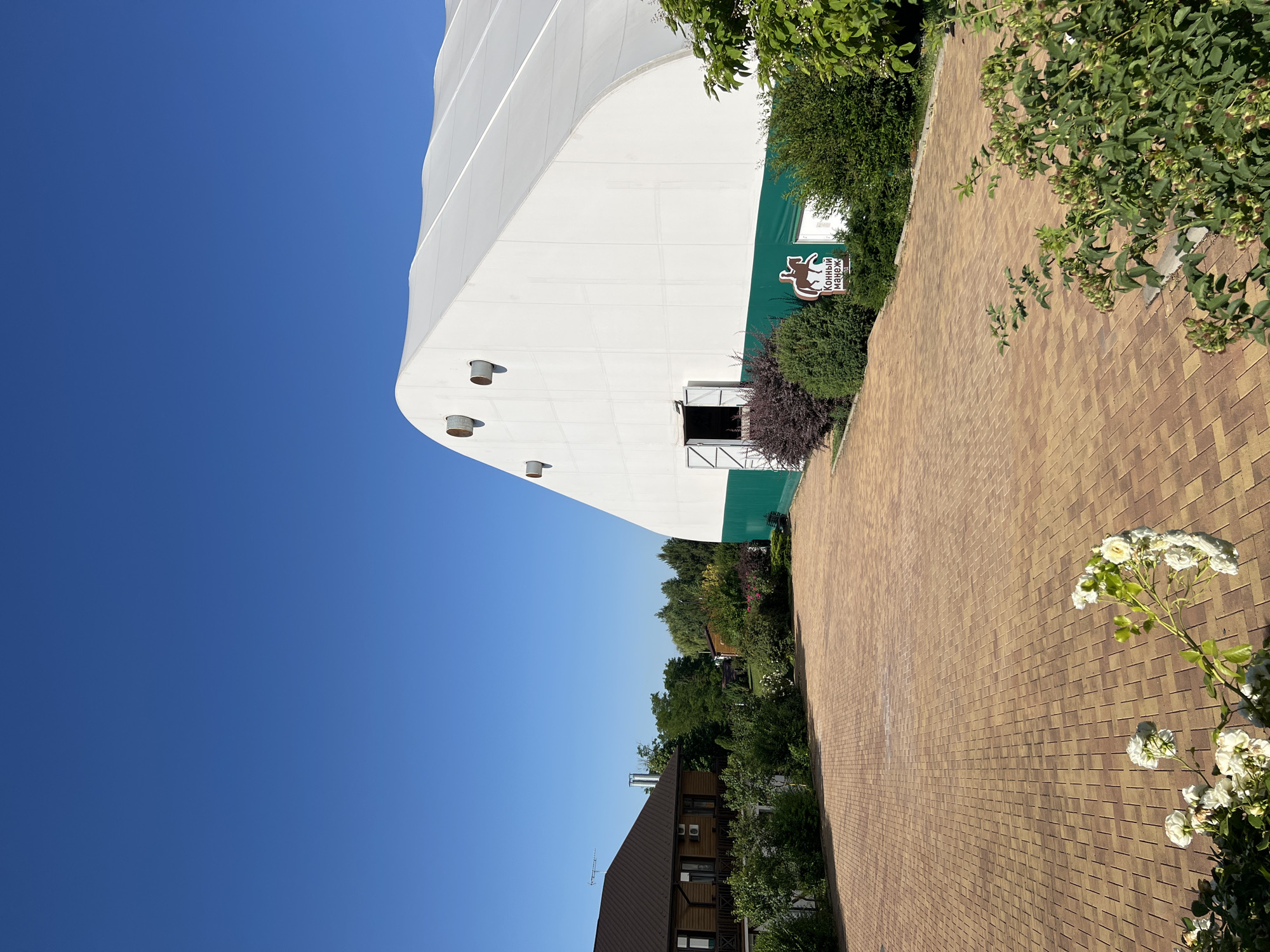 The image captures a sunlit outdoor scene at a facility in Plastunivska, Russia, under a clear blue sky. In the foreground, a paved area made of interlocking reddish-brown bricks leads towards a large, white, arched tent-like structure. This structure, with a green band around its base, has an open doorway. To the left of the paved area, a two-story wooden house with a dark brown roof is visible, surrounded by lush green trees and bushes. A sign with a silhouette of a horse and rider, and text in Russian, is placed in front of the tent. The middle ground and background are filled with dense green foliage, indicating a natural or park-like environment. White flowers are blooming in the lower left corner of the frame.