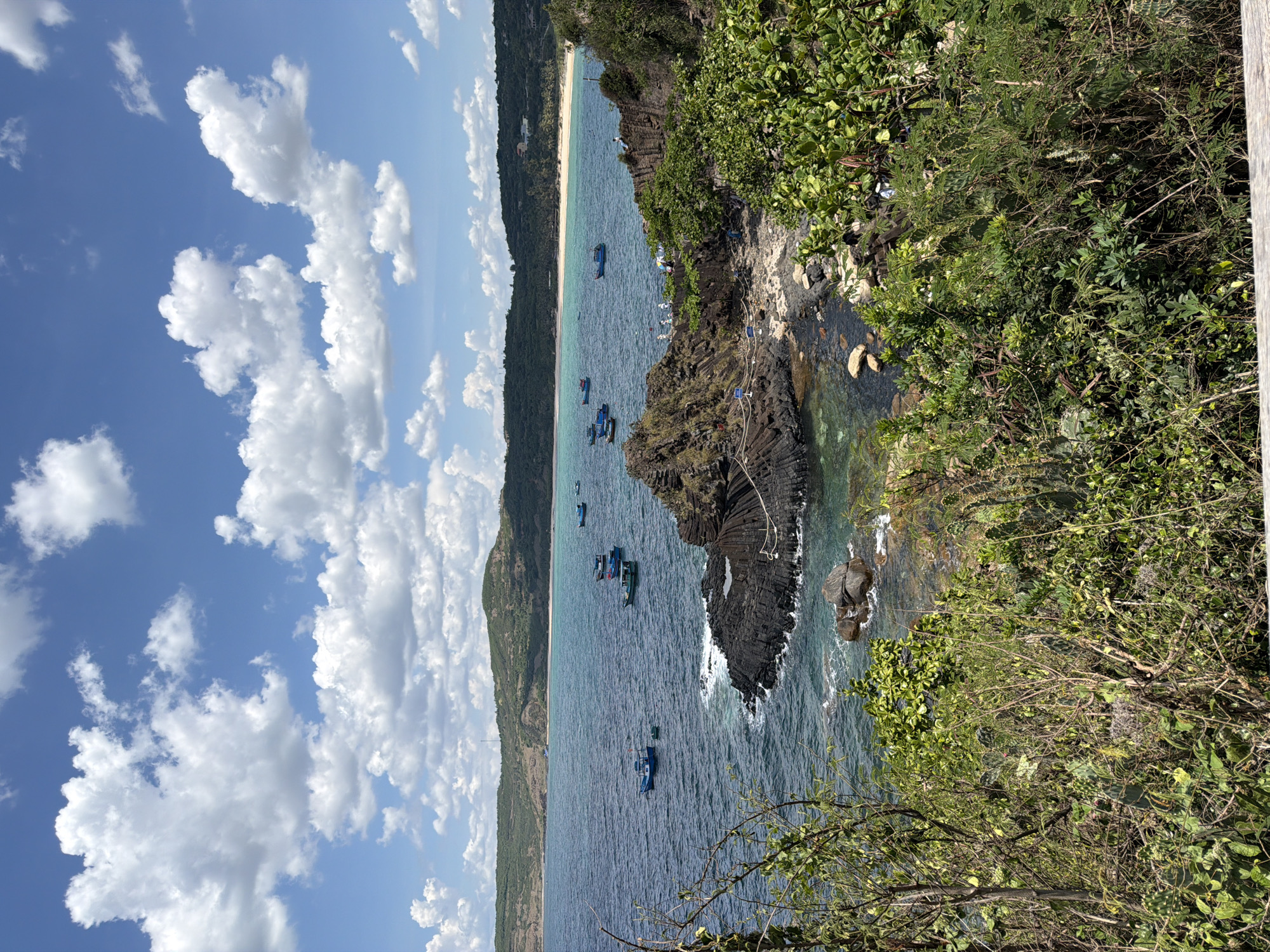 A bright, sunny day with a blue sky filled with puffy white clouds is captured in this medium-angle shot of a coastal landscape. The foreground is dominated by lush green foliage and shrubbery, some of which is prickly, obscuring a wooden railing. Beyond the vegetation, a choppy, deep blue ocean stretches out, dotted with several small, blue fishing boats. To the right, a rocky outcrop of dark, basaltic columns juts into the sea, with a few people visible on its upper surface. The middle ground features a white sandy beach backed by rolling, green hills that extend into the distance.