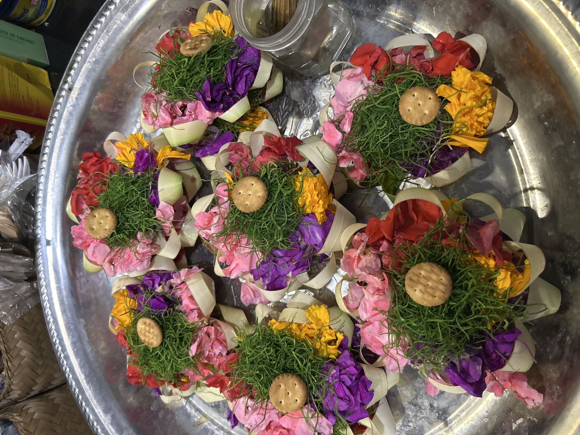 The image displays a collection of offerings, likely for a religious or spiritual ceremony, arranged on a large, round, metallic tray. The offerings are elaborately constructed, each featuring a base of what appears to be woven palm or banana leaves, forming cone-like structures. These cones are filled with vibrant flower petals in shades of pink, red, purple, and yellow, interspersed with a dense green material that resembles fine grass or moss. Topping each offering is a small, round, light-brown cracker with visible perforations.
There are at least six such offerings visible in the frame, clustered together on the tray. The metallic tray has a slightly textured surface and a raised rim. In the background, some items are partially visible, including a clear plastic container holding small wooden sticks, suggesting the offerings might be for an incense-related ritual. There are also glimpses of packaging and woven baskets, hinting at a market or roadside stall setting, consistent with the provided location context of Sukawati, Indonesia.
The overall impression is one of meticulous preparation and vibrant color. The lighting suggests it is daytime, though no specific weather conditions can be determined. The scene evokes a sense of cultural tradition and reverence. A partial label on a green box in the upper left corner reads "ICE IN SACHET."