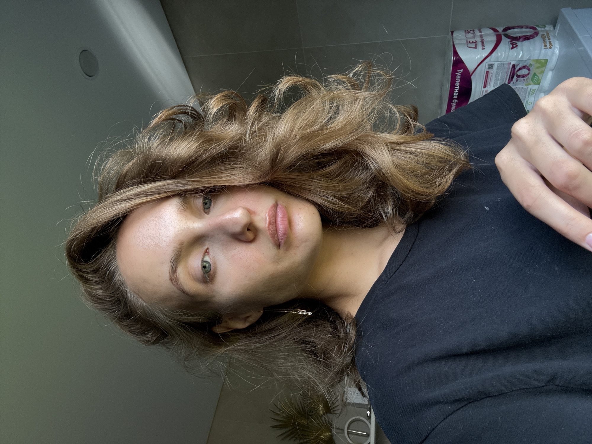 A medium shot shows a young woman with medium-length, wavy brown hair and fair skin. She is looking directly at the camera with a neutral expression. Her eyes are light blue-green, and her lips are slightly parted. She is wearing a black t-shirt. A gold earring with small, clear stones hangs from her left ear. Her right hand is in the lower right corner of the frame, fingers slightly curled.
The background appears to be a bathroom. To the left, a shelf with decorative items, including a gold fan-like object and a clock, is visible. To the right, a stack of toilet paper rolls is on a shelf or surface. The walls are tiled in a neutral tone. The lighting suggests the image was taken indoors during the day, possibly near a window, casting some shadows.
The image does not contain any explicit location markers for the city of Подшивалово, Russia.