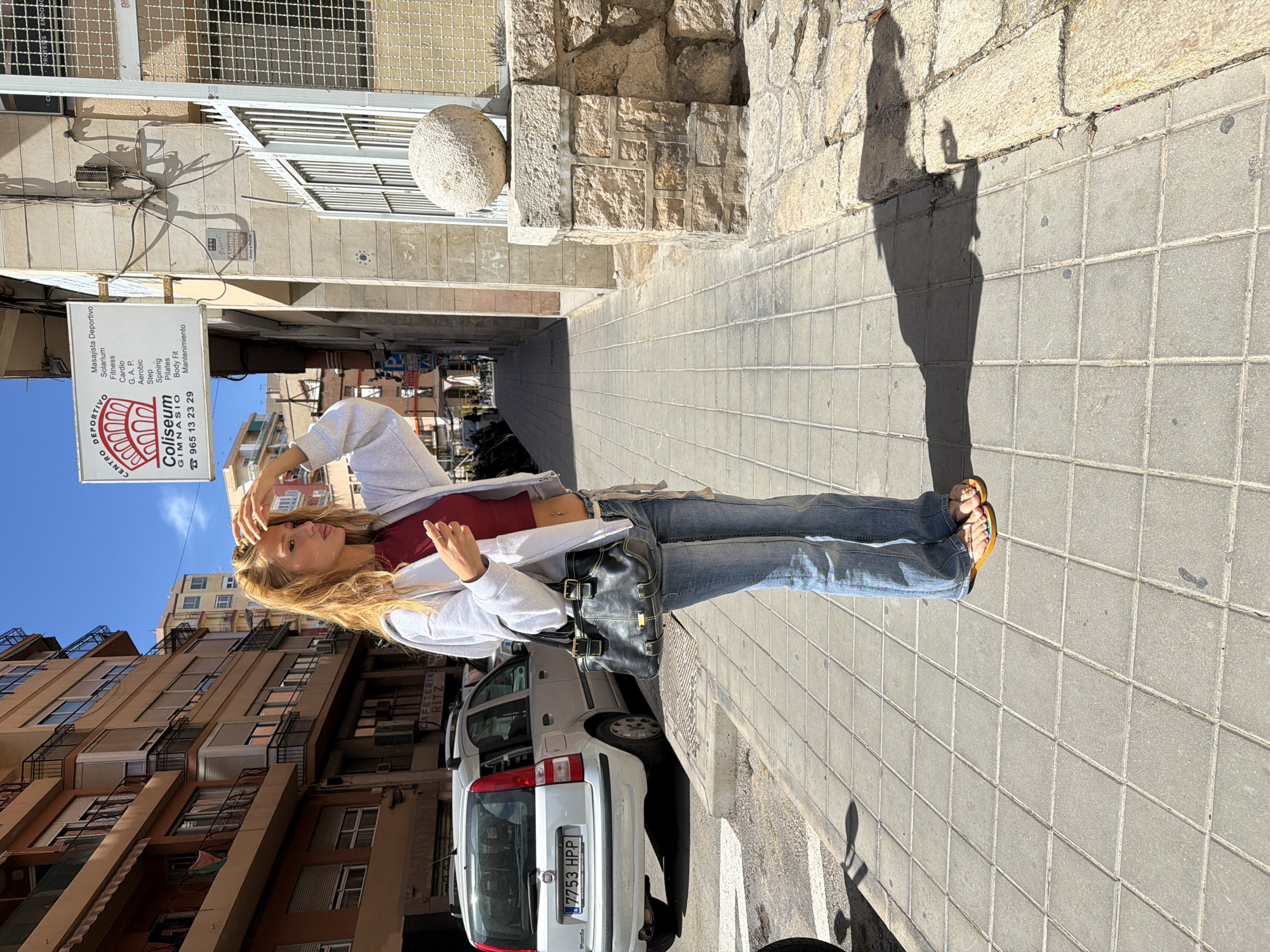 The image depicts a young woman standing on a tiled sidewalk in Alicante, Spain. She has long blonde hair and is wearing a red top, a grey hoodie, and blue jeans. She has a black bag slung over her shoulder and is wearing sandals. She is holding her hand up to her head, possibly shielding her eyes from the sun.
The background features buildings with apartments and a white car parked on the street. Above her head, there is a sign for "Coliseum Gimnasio" as well as another for "CAFETERIA RITZ". The sky is blue and sunny. The scene is a street or alleyway.