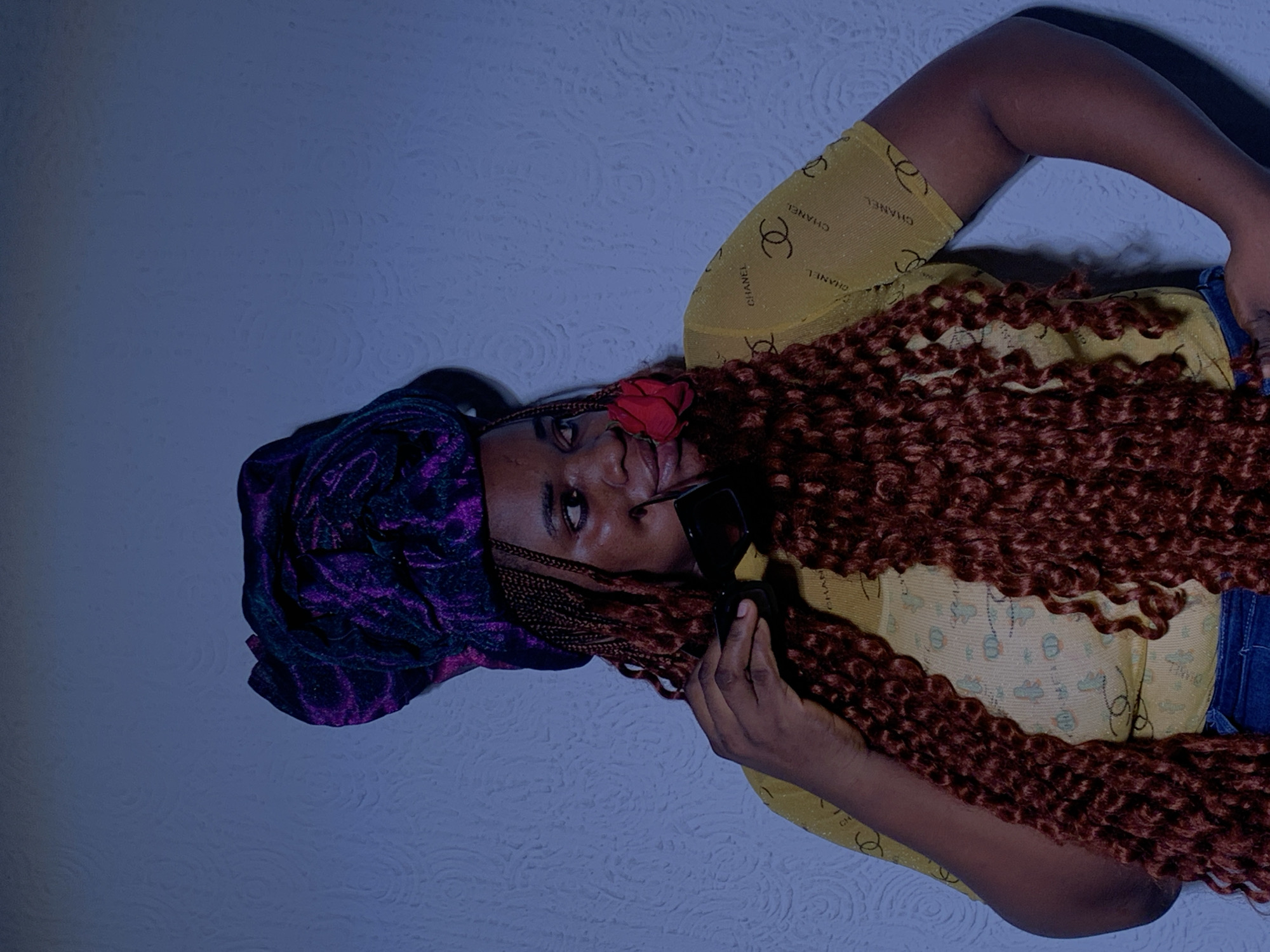 An overhead shot captures a woman with dark skin, positioned diagonally against a textured, light blue-grey wall. She looks directly at the camera with a neutral expression. Her head, adorned with a dark purple, possibly velvet, patterned headwrap, is situated towards the upper left of the frame. A small red flower is tucked into her hair above her right temple.
Her hair consists of long, dark reddish-brown extensions, styled in tight coils or braids, which cascade across her chest and right shoulder. She wears a translucent yellow long-sleeved top, visibly patterned with repeating black "CHANEL" logos. Her right hand, holding a pair of black sunglasses, is raised near her face. A sliver of blue denim, likely from jeans, is visible around her waist. The background is a plain wall with a subtle textured pattern. The lighting suggests an indoor setting, with soft shadows creating depth. The scene is set in Barakin Akawo, Nigeria.