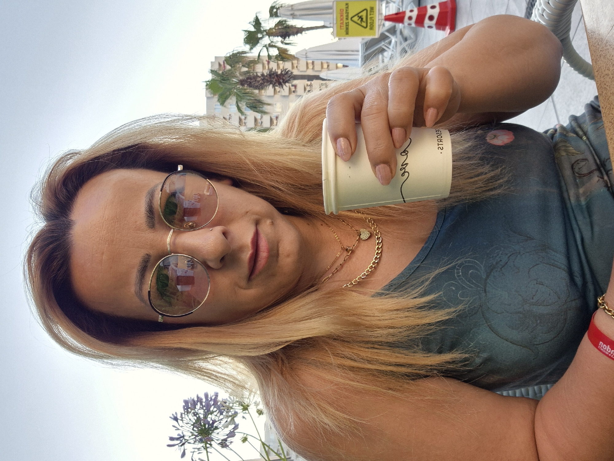 A woman with blonde hair and sunglasses is taking a selfie while holding a cup. She is wearing a teal colored top with a patterned design and a gold chain necklace with a heart pendant. A red wristband with text is visible on her right wrist. In the background, there are palm trees and a building. A yellow sign with black text and a warning symbol is also visible. A purple flower is in the foreground. The sky is overcast. The scene appears to be outdoors, possibly a cafe or resort. The lighting suggests it is daytime.