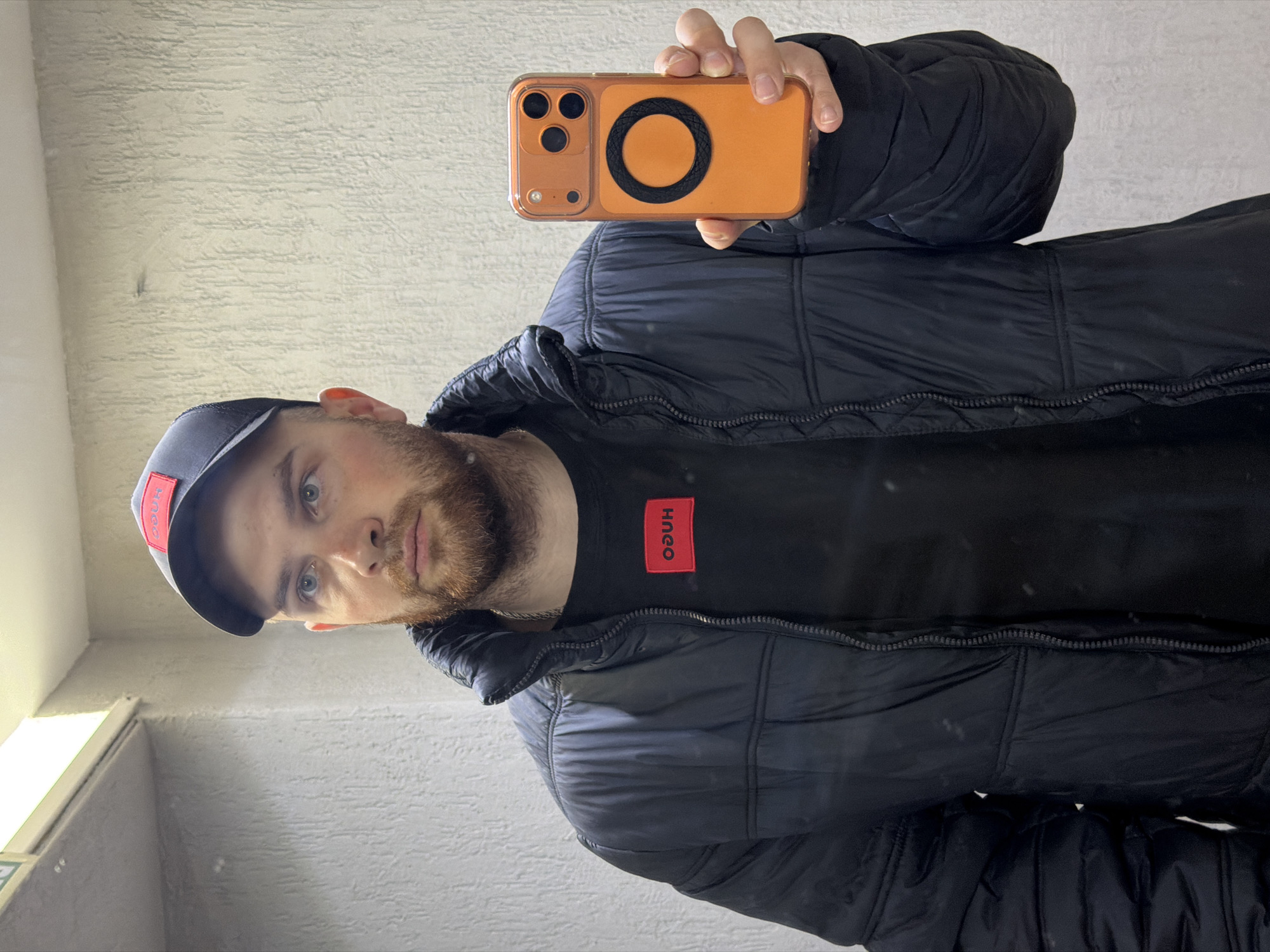 A man is taking a selfie in front of a textured light-colored wall. He is wearing a black puffer jacket, a black t-shirt with a red "HUGO" logo on the chest, and a black baseball cap with a red "HUGO" logo on the front. He has a beard and is holding an orange phone with a black circular object on the back. The location is the city of Chaikovsky, Russia.