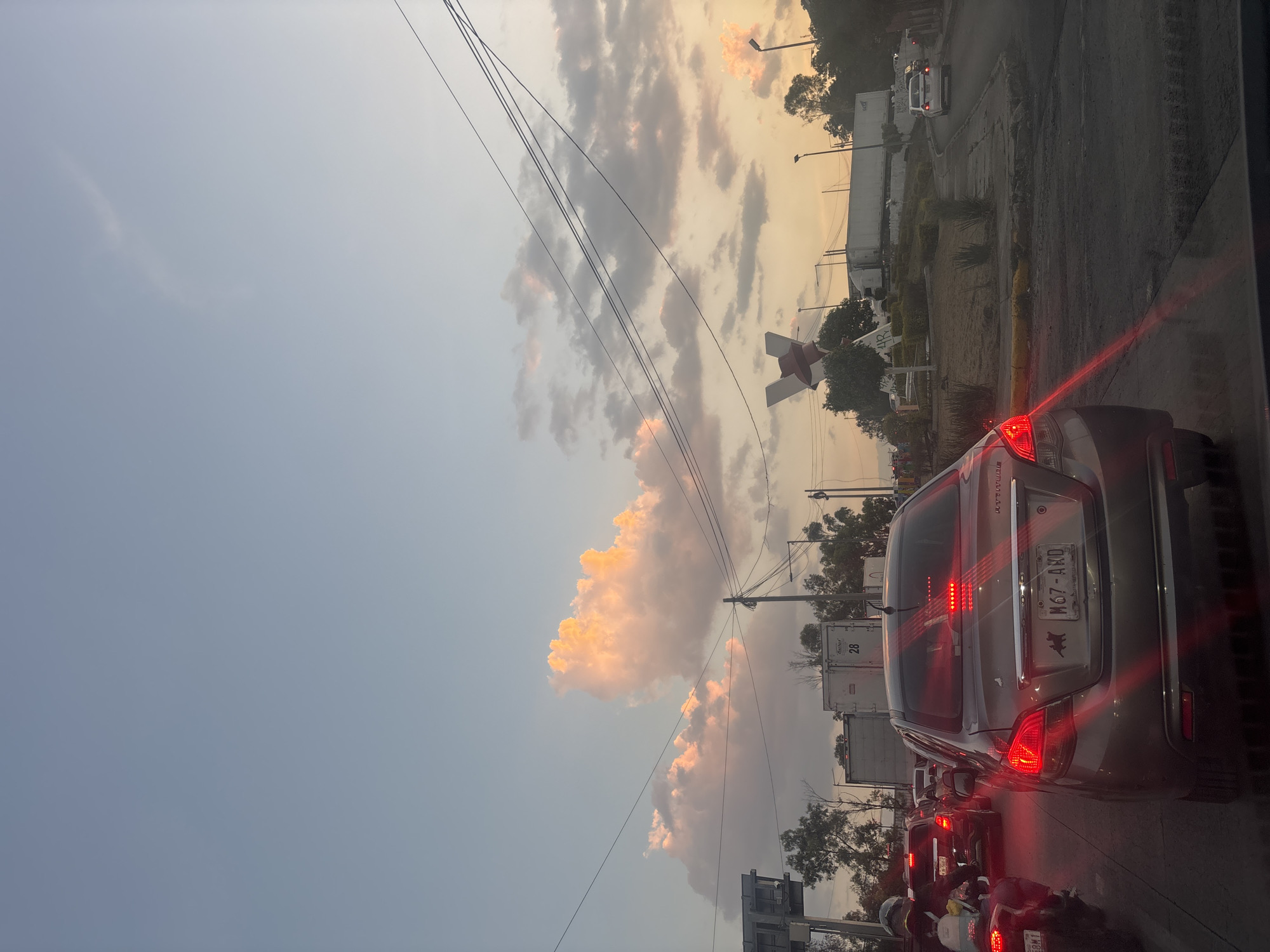 The image shows a congested traffic scene in Buenavista, Mexico, during what appears to be late afternoon or early evening, based on the sky's color and the lighting. Multiple vehicles are stopped or moving slowly, including a gray car directly in front of the camera with its brake lights illuminated. The license plate is visible and reads "M67-AND." Behind it, other cars and motorcycles are discernible in the traffic. Overhead power lines stretch across the sky, and in the background, there are trees, buildings, and possibly trucks or trailers. A large, red and white X structure is visible, potentially a landmark or advertisement. The sky is cloudy, with some clouds reflecting a reddish-golden hue from the setting sun. The environment suggests an urban area, experiencing typical rush hour traffic.