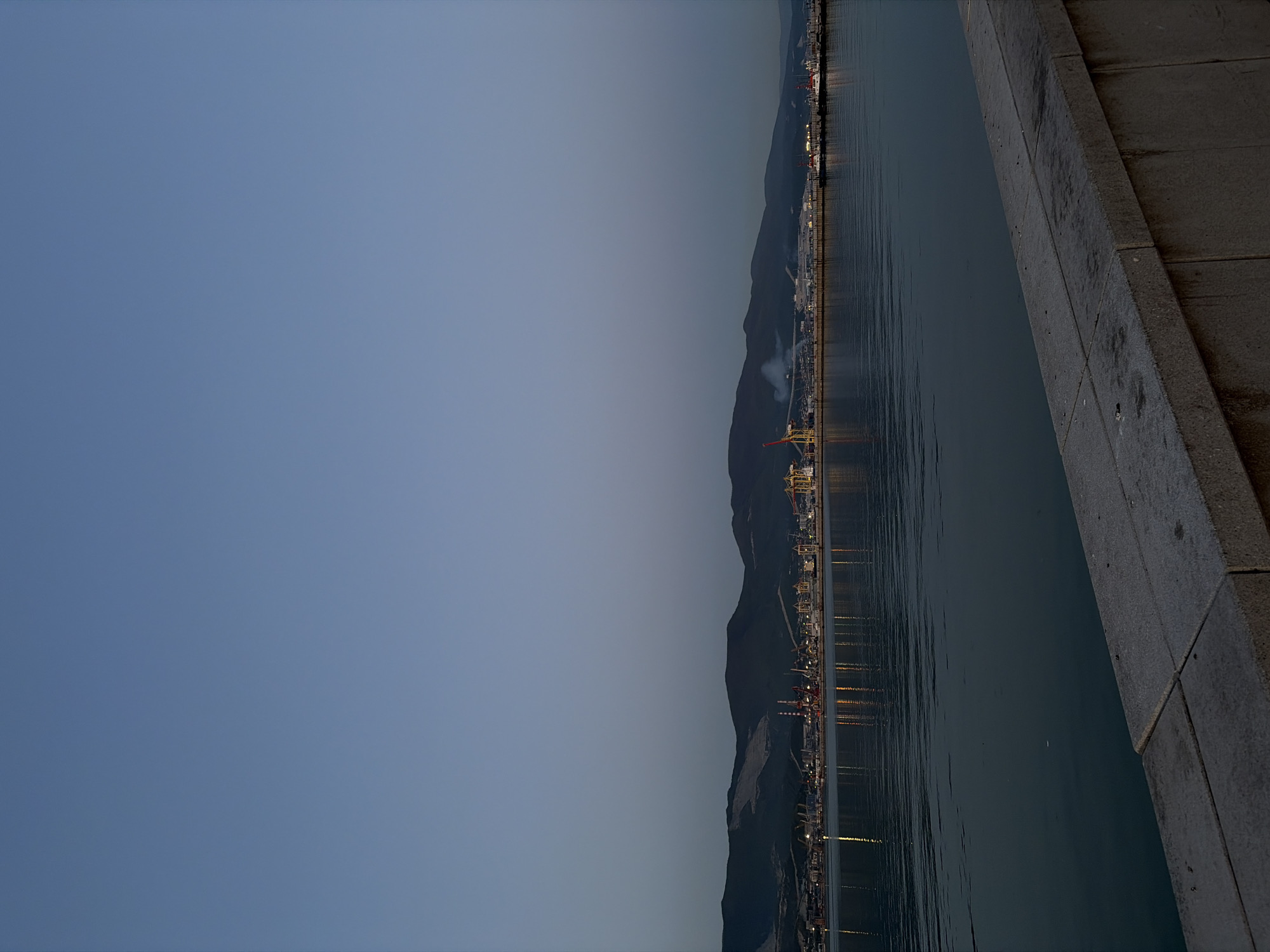 The image depicts a waterfront view in Novorossiysk, Russia. The right side of the image shows a concrete embankment, alongside the dark water of the bay. Across the water, a stretch of land is visible, featuring the city's industrial zone with port facilities, mountains in the background, and a clear sky. Lights from the city and port reflect on the water's surface. The sky is light.