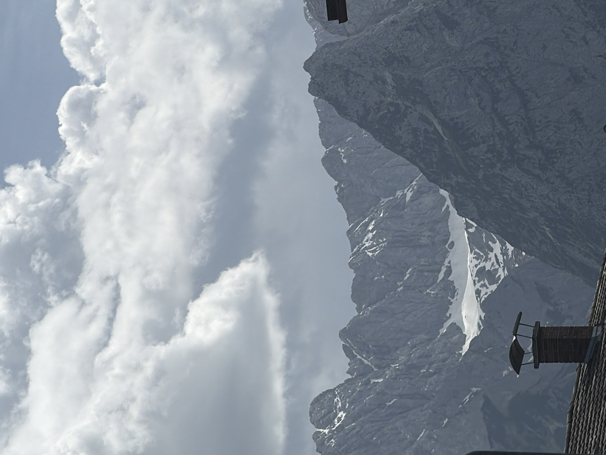 The image shows a mountain landscape with a cloudy sky. The mountains are rocky and rugged, with patches of snow visible. A building with a chimney is partially visible on the right side of the image. The scene is likely located in or near Garmisch-Partenkirchen, Germany, given the alpine environment.