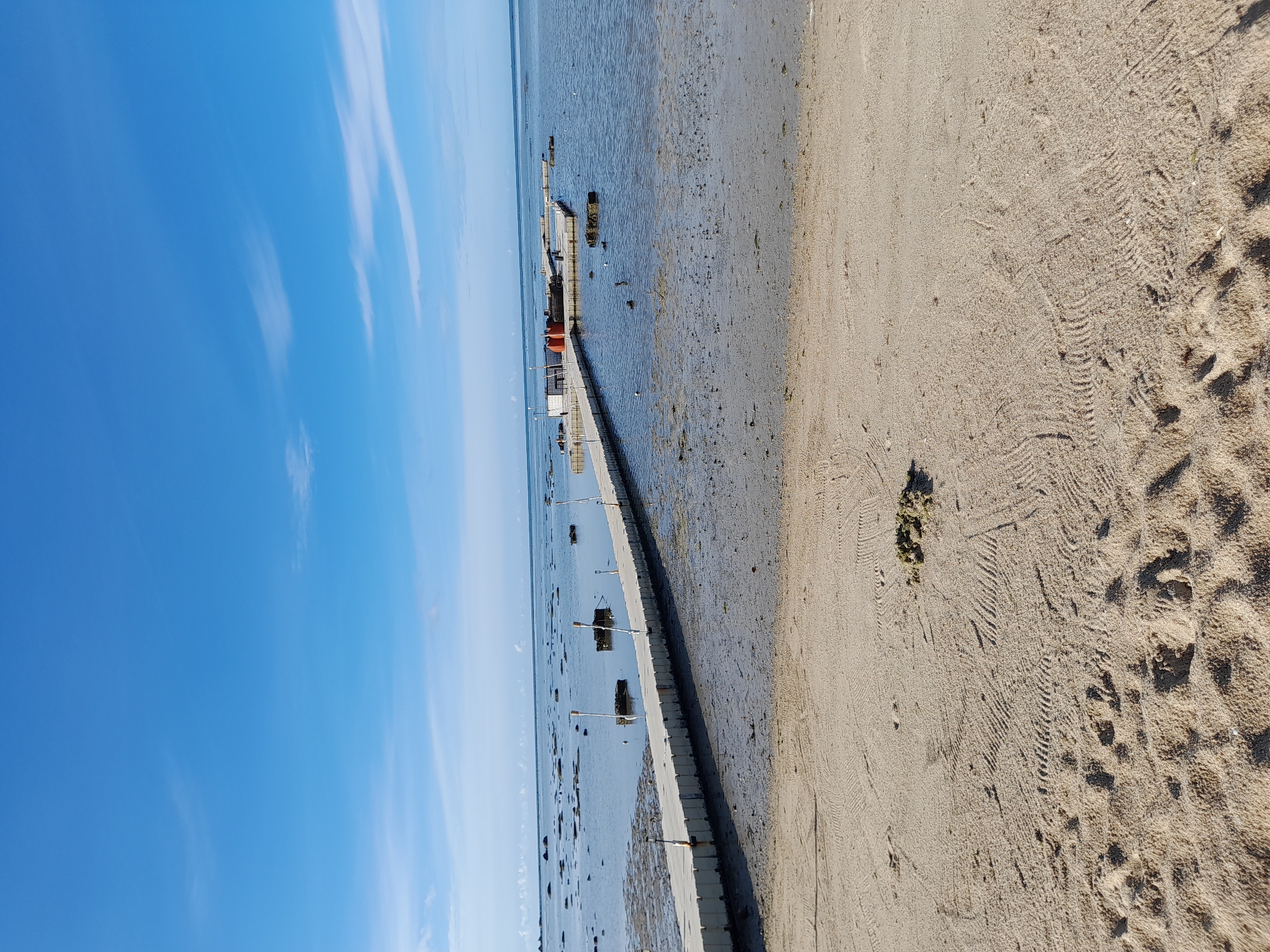 A long, white floating dock extends from a sandy beach into a shallow body of water under a clear blue sky with wispy white clouds. The dock is made of interconnected modular plastic sections and has small poles with lights spaced along its length. In the distance, the water is calm and reflects the sky, with scattered dark rocks and patches of seaweed visible beneath the surface. A larger floating structure, possibly a small platform or raft with some objects on it, is situated further out in the water. The sandy beach in the foreground shows tire tracks and footprints, indicating recent activity. The overall scene depicts a serene coastal environment, likely during daytime.