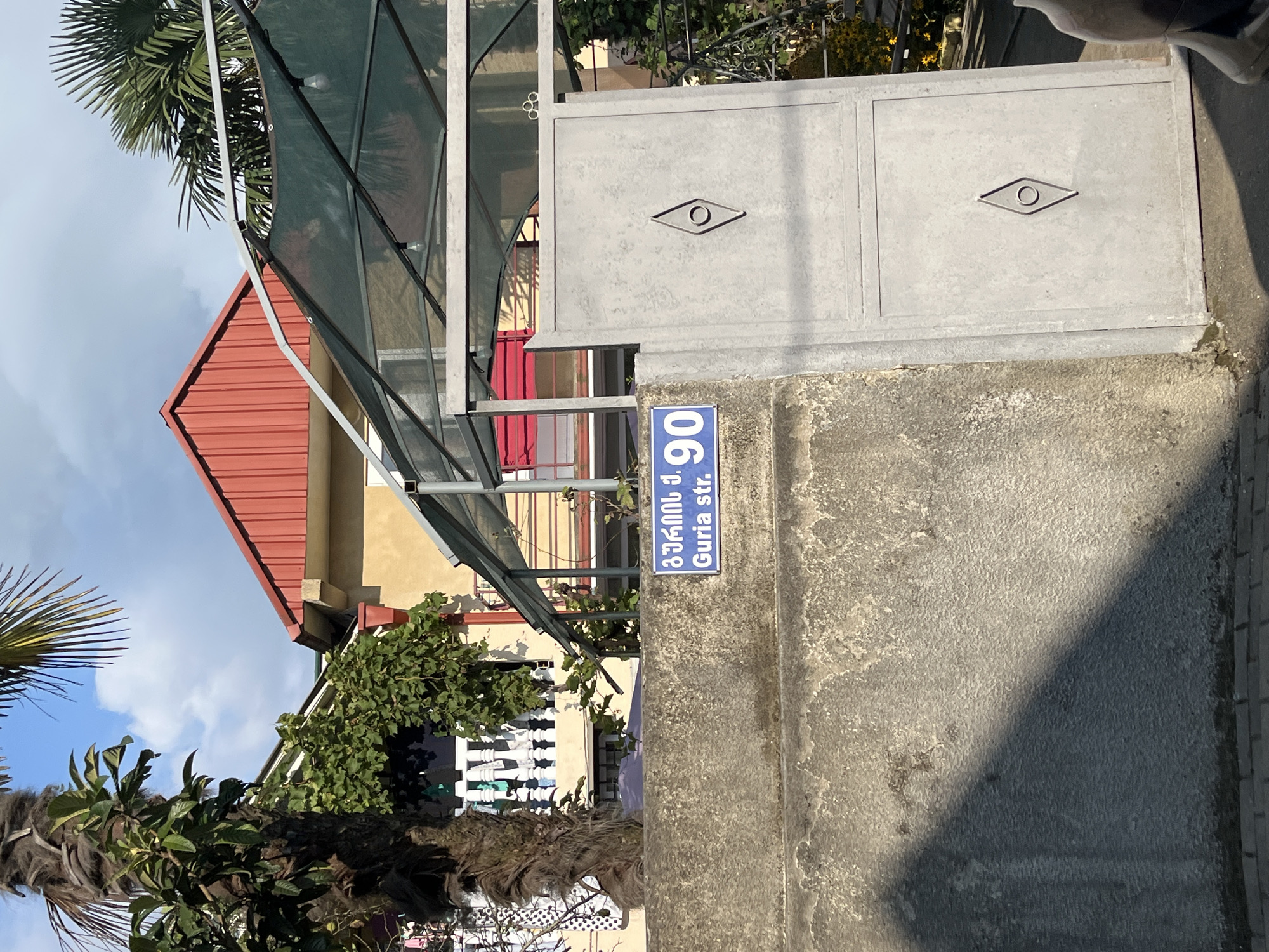 The image shows a street sign in Ureki, Georgia, with Georgian script and the text "Guria str. 90". The sign is blue with white text and is attached to a concrete wall. Behind the wall, there is a residential building with a red roof, a balcony with white railings, and a large palm tree. A metal gate with a diamond-shaped ornament is to the right of the sign. The environment appears to be outdoors on a sunny day, with palm trees and foliage suggesting a warm climate. The area seems to be a residential street.