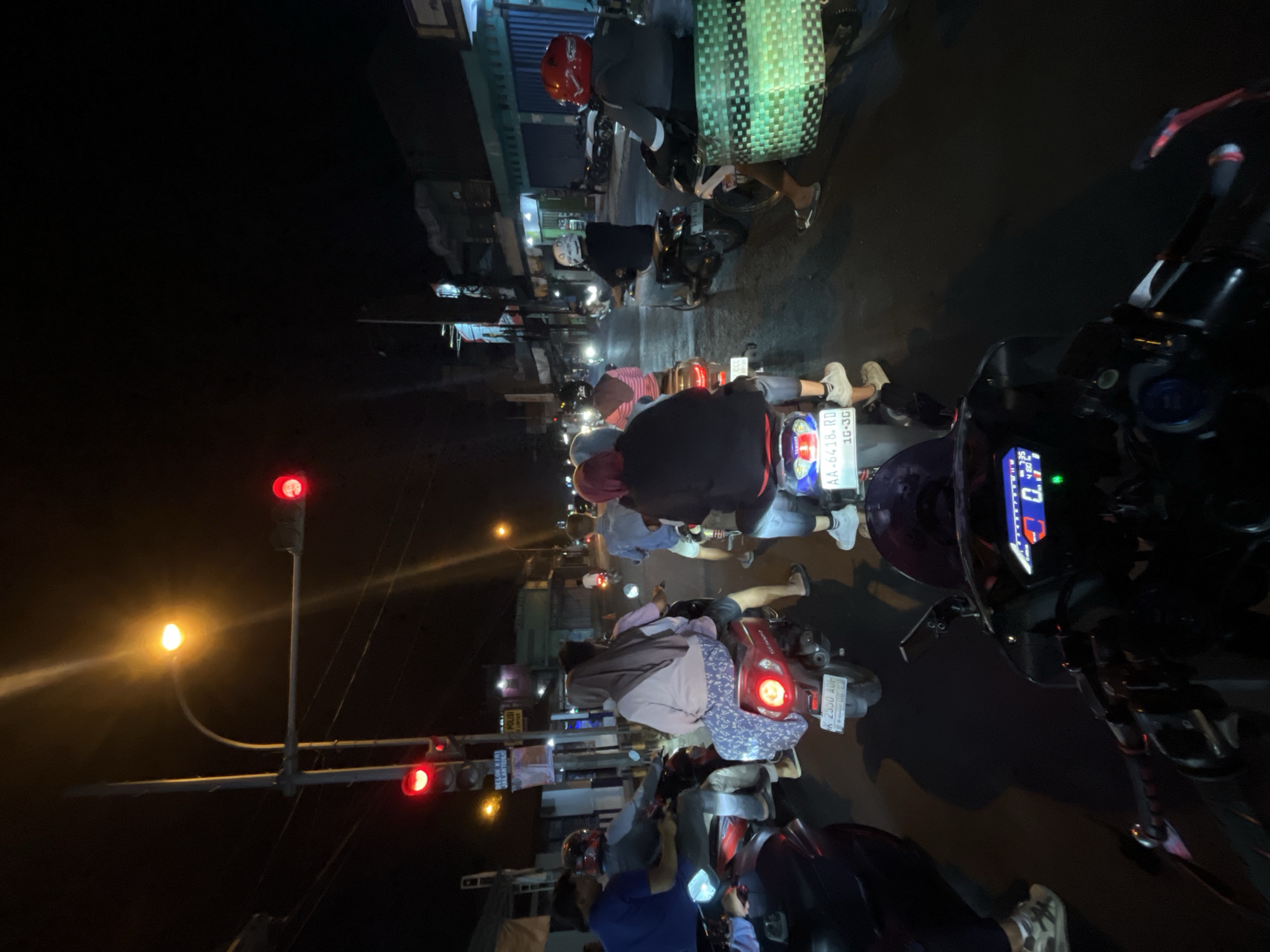 A nighttime street scene in Kalinyamatan, Indonesia, depicts a traffic jam on a paved road. Several motorbikes are stopped at a traffic light, with riders and passengers visible. The traffic light is red. Buildings line the street, some with lights on. The overall lighting is dim, characteristic of nighttime.
